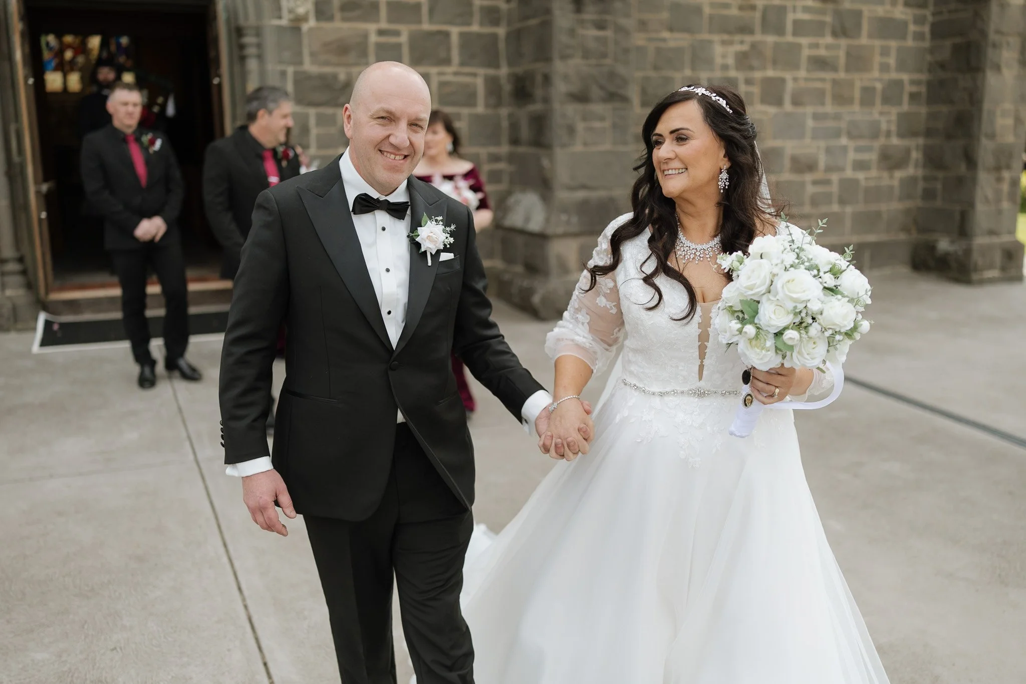 A newlywed couple, a man in a black tuxedo with a bow tie and a woman in a white wedding gown holding a bouquet, walking hand in hand outside a church with a stone wall. The bride is smiling and looking at the groom. In the background, two men and a 