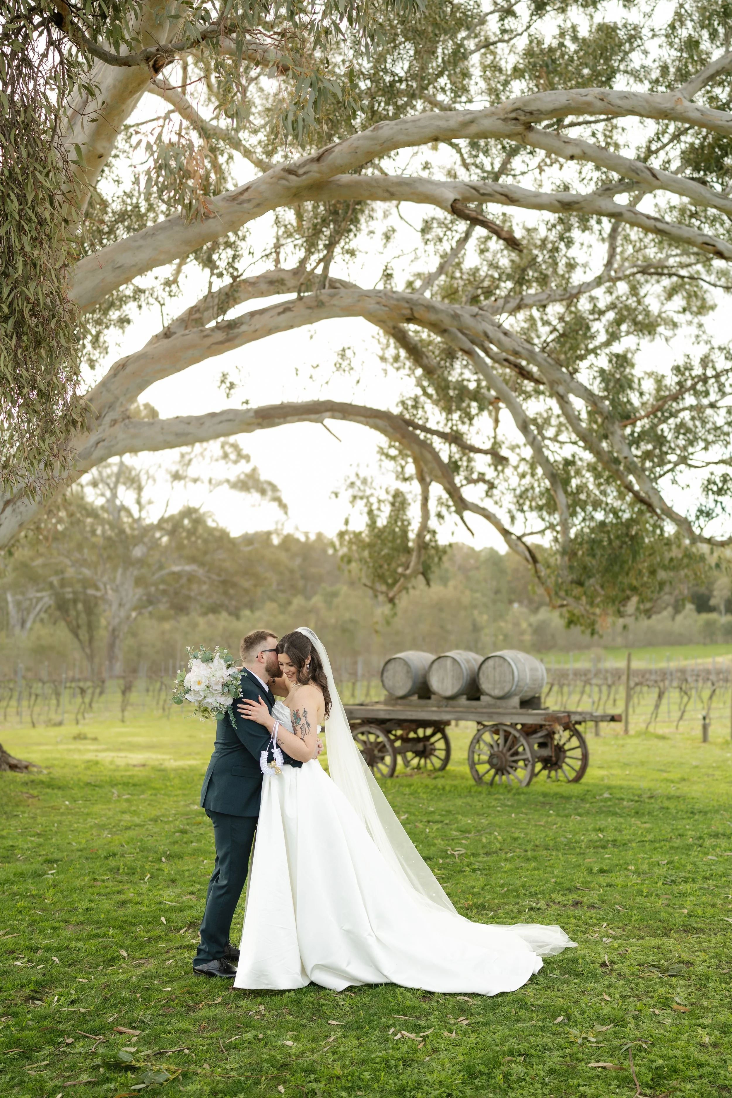 A newlywed couple embrace under a large tree in a vineyard, with the groom in a black suit holding a bouquet of white flowers, and the bride in a white wedding gown with a long veil, standing on green grass with rolling hills and a rustic wooden wago