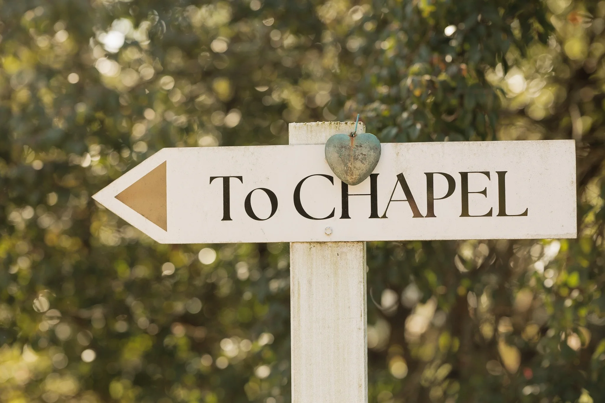 A white directional sign pointing left with the word 'TO CHAPEL' and a rusty heart-shaped decoration at the top, set against a backdrop of green foliage with sunlight filtering through.