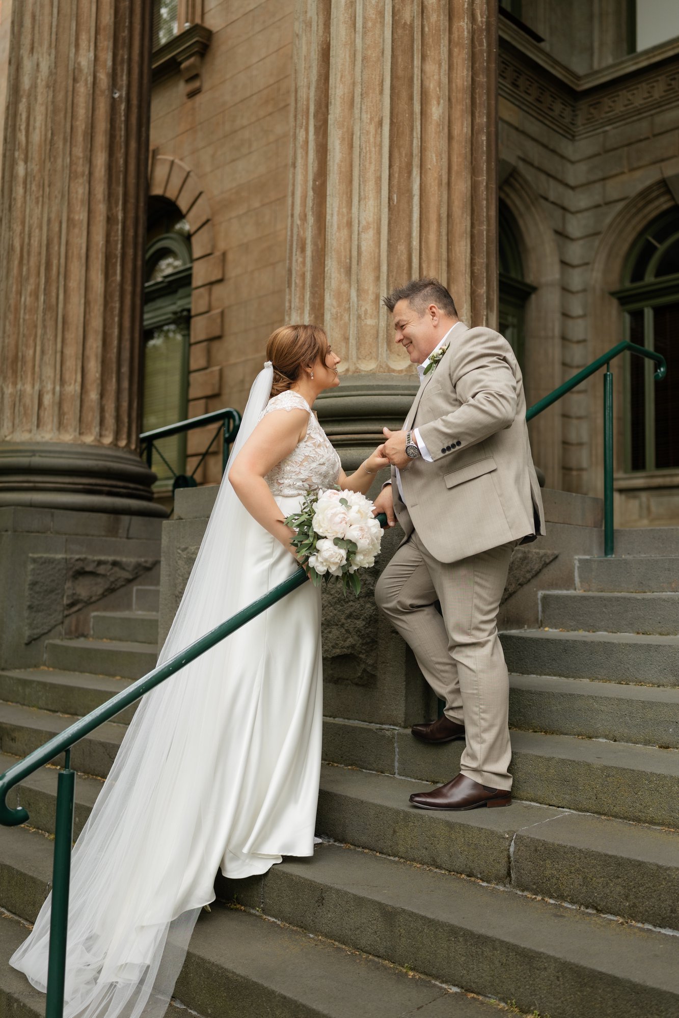 A bride and groom standing on outdoor stairs in front of a historic building, smiling and gazing into each other's eyes, holding hands, with the bride holding a bouquet of white flowers.