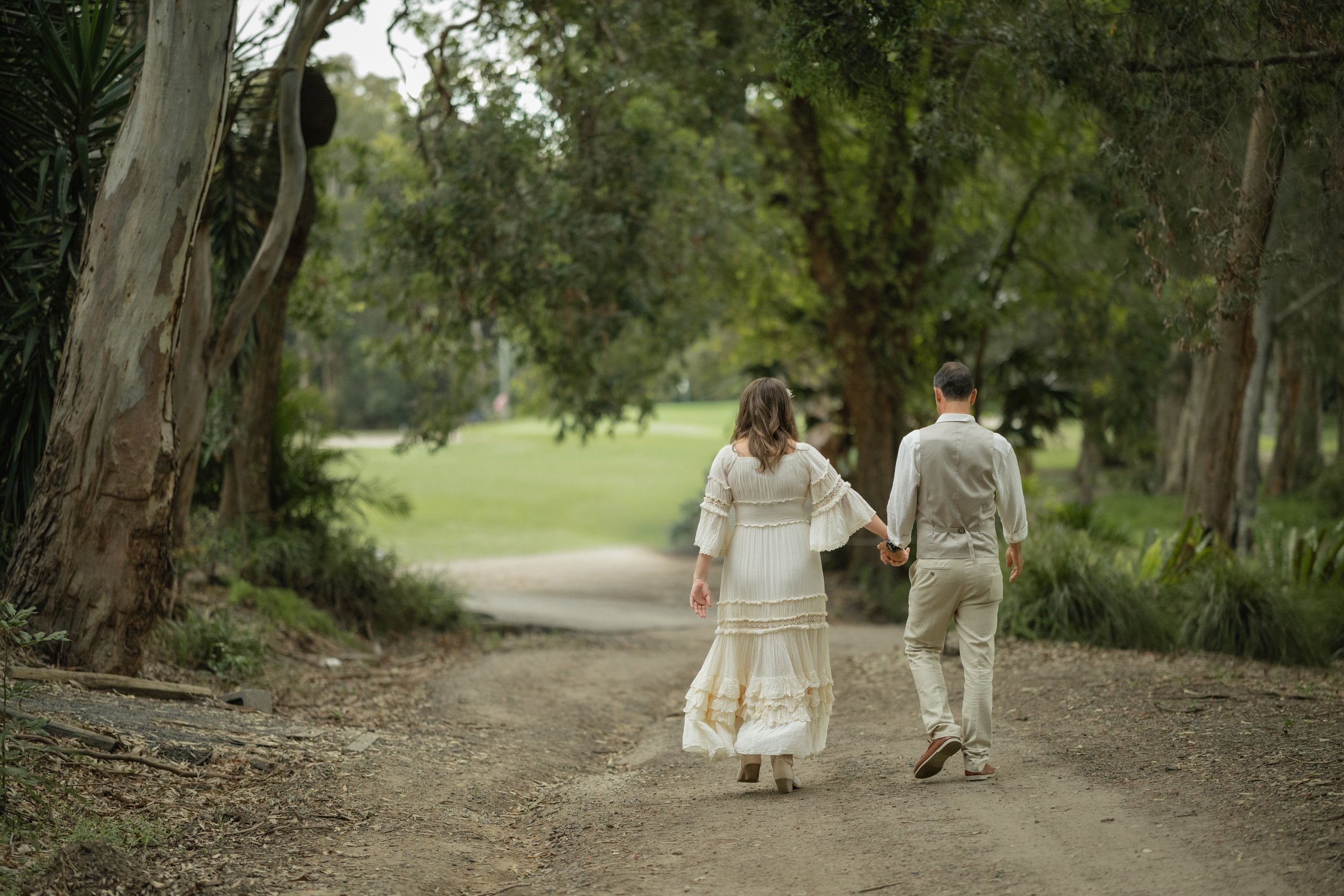 A couple in vintage attire walking hand in hand down a wooded dirt path, surrounded by trees and greenery.