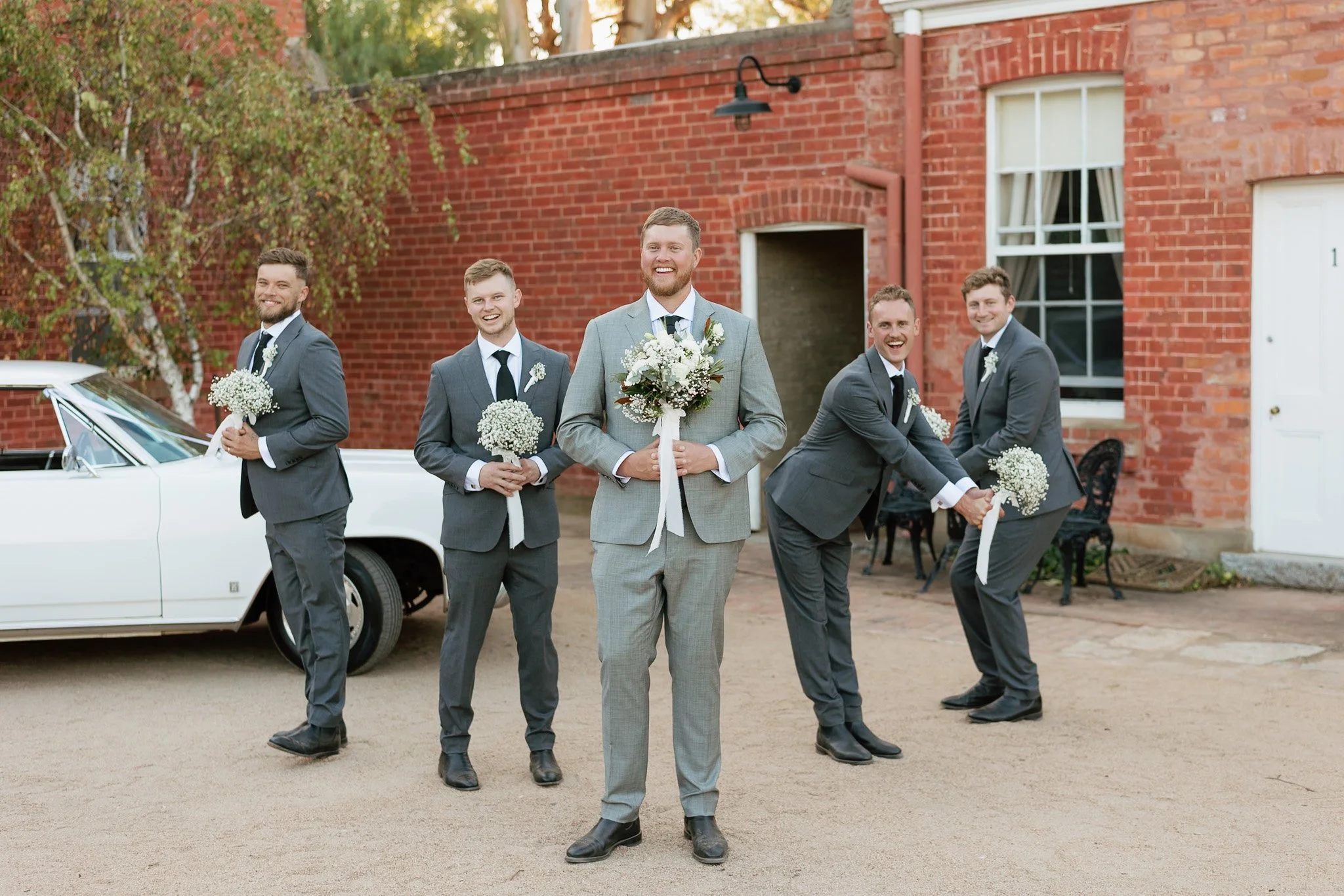 A group of five men dressed in suits holding small bouquets of flowers, standing outdoors in front of a red brick building, smiling and posing for a photo during a wedding.