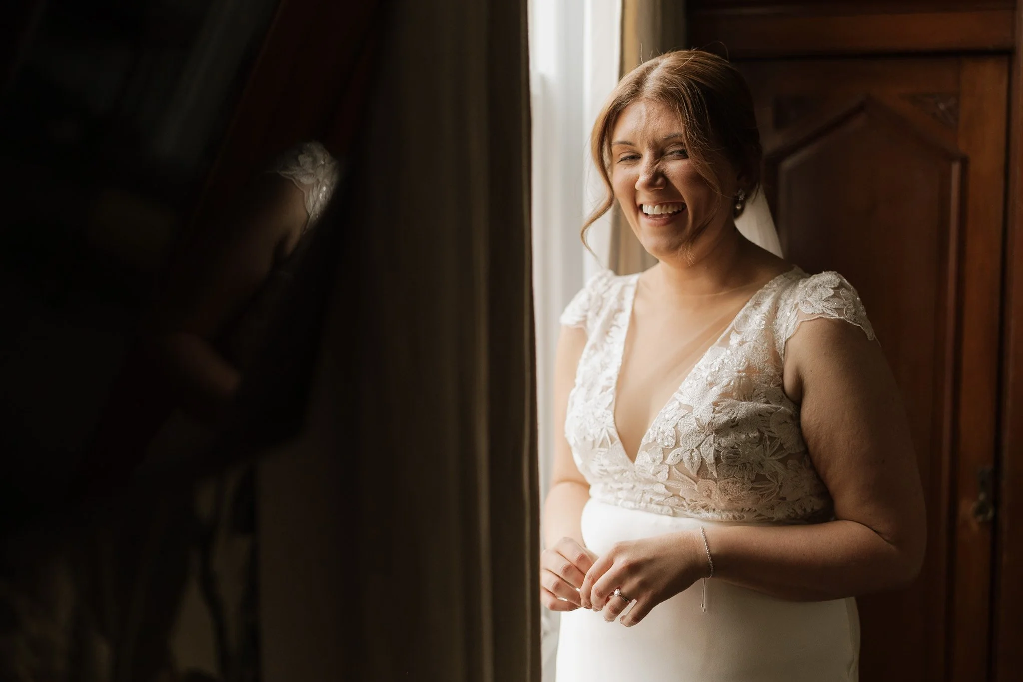 A woman in an elegant wedding dress standing by a window with natural light, smiling softly.