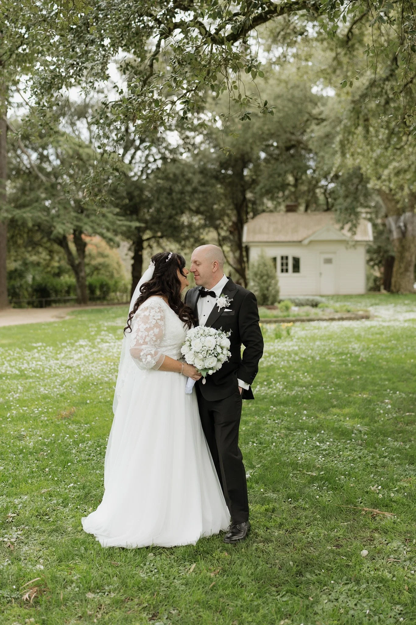 A bride and groom standing close together outdoors on a grassy lawn with trees and a small white house in the background, sharing a tender moment on their wedding day.
