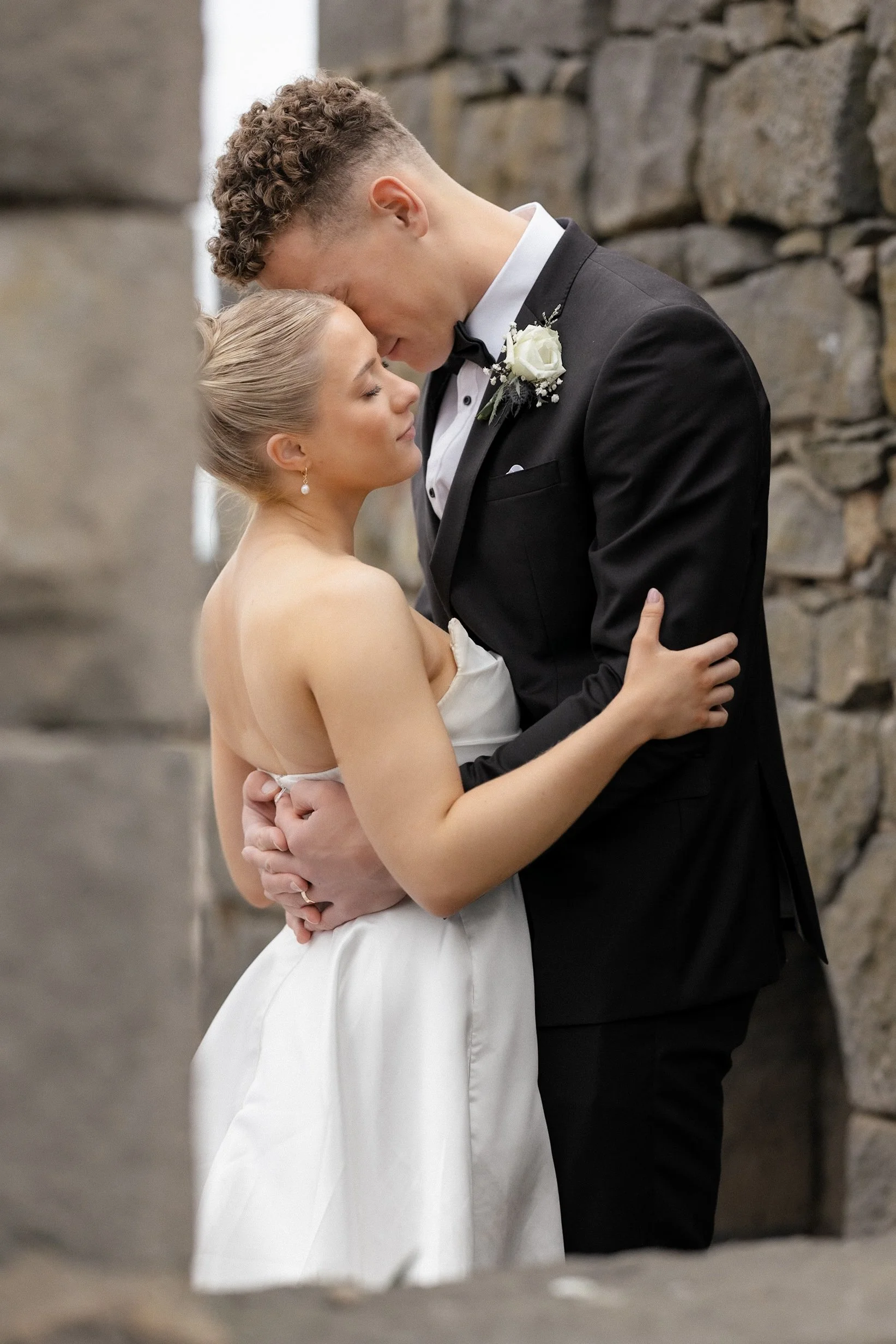 A bride and groom sharing a romantic moment outdoors, embracing each other with their foreheads touching, near a stone wall.