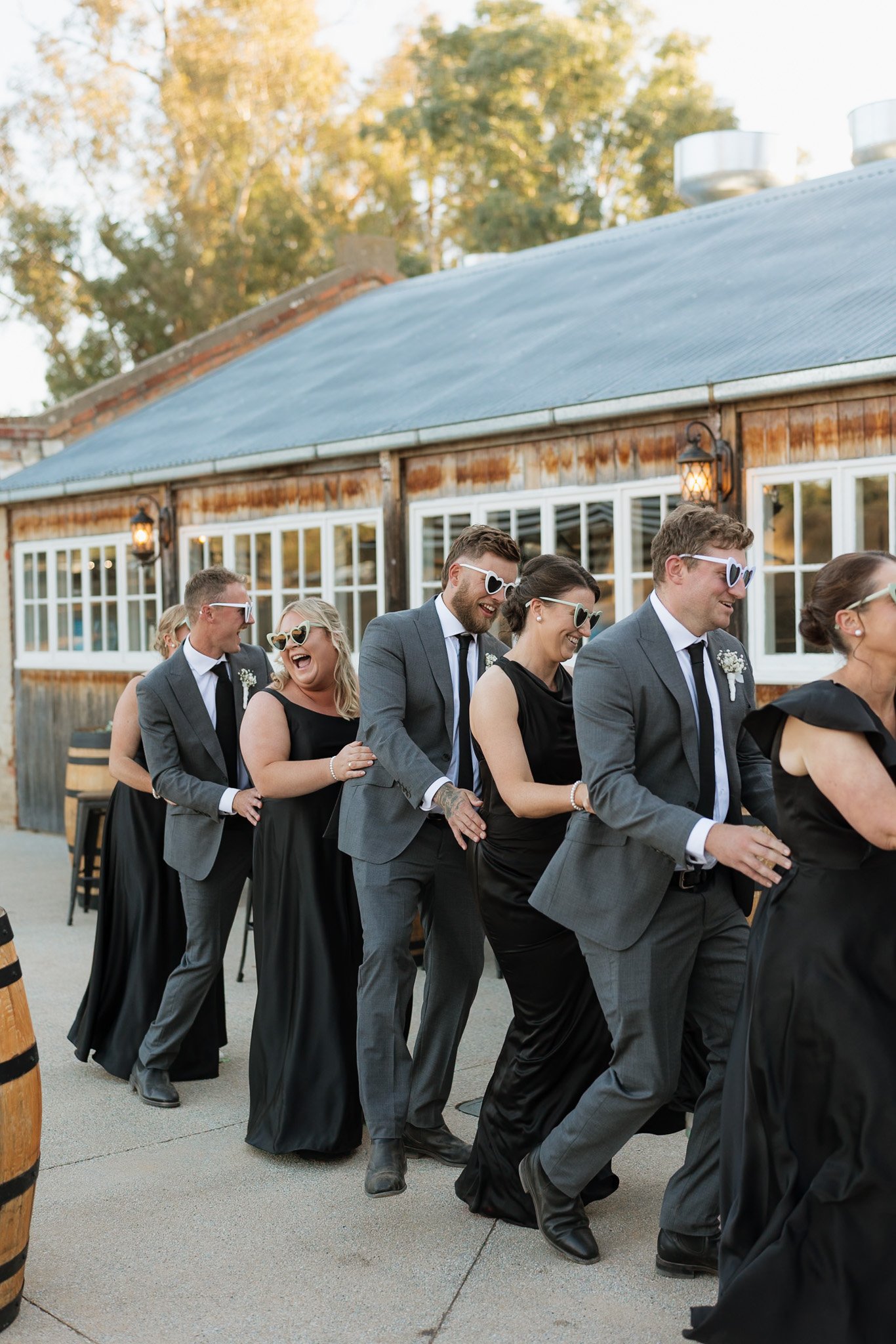 A group of people dressed in formal black-tie attire, including men in suits and women in black dresses, are standing in a line outdoors. They are all wearing heart-shaped sunglasses and appear to be enjoying a social event, possibly a wedding, in fr