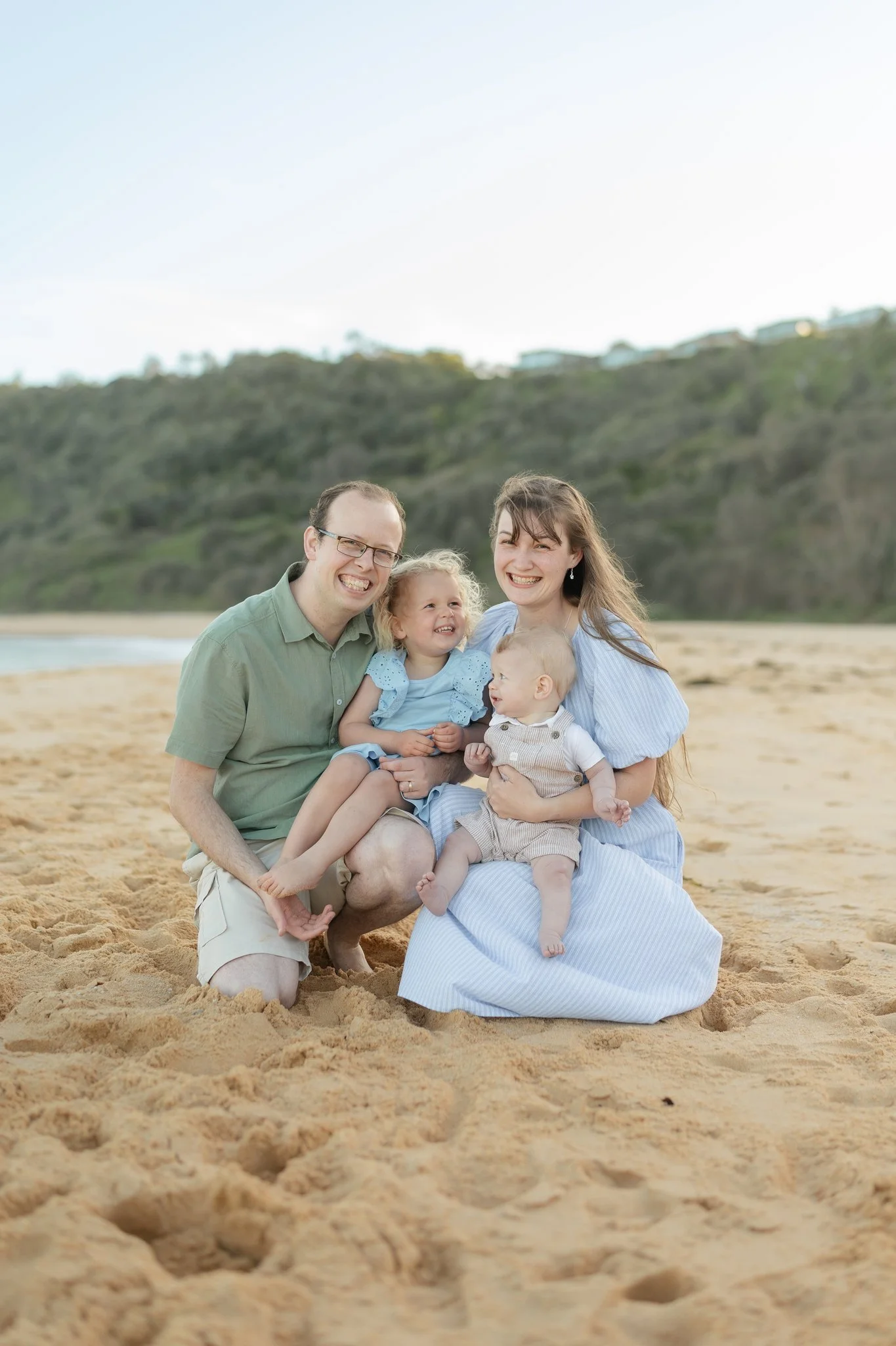 A happy family of four sitting together on a sandy beach with green hills in the background, smiling at the camera.