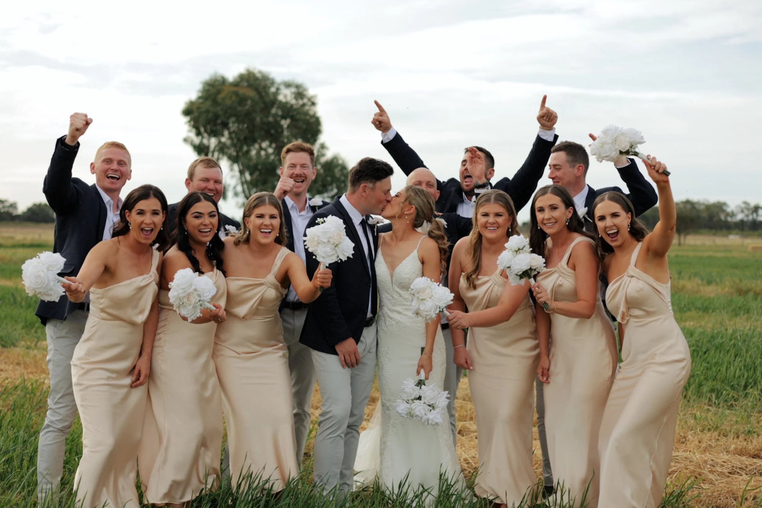 A wedding party of eleven people, including the bride and groom, celebrating outdoors in a field. The bride and groom are kissing, holding bouquets of white flowers. The bridesmaids are in light beige dresses holding bouquets, and the groomsmen are in suits. The group is smiling, cheering, and showing excitement against a backdrop of grass, trees, and a cloudy sky.