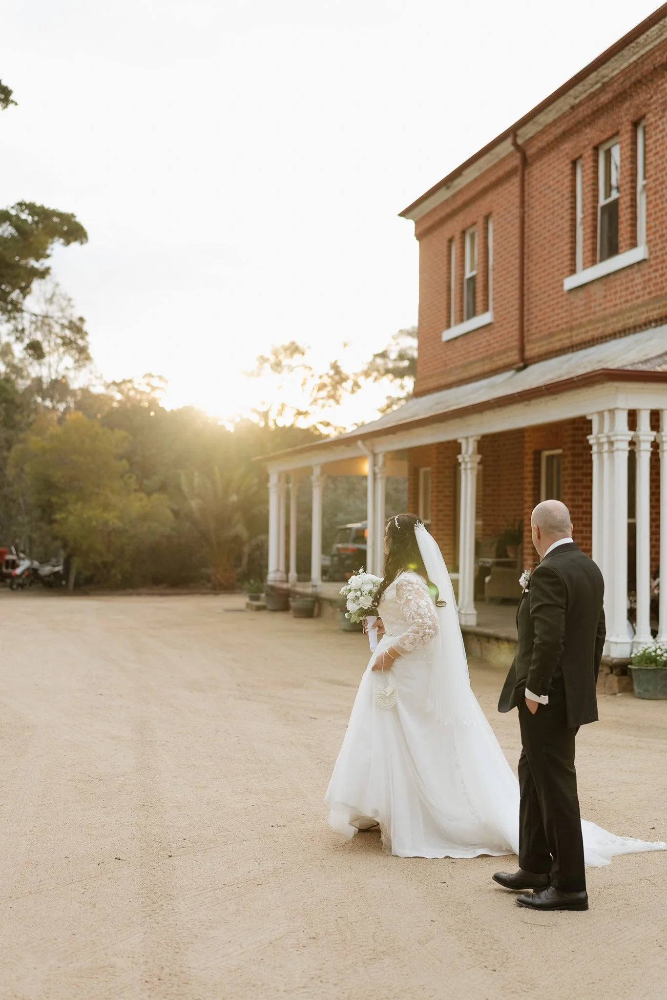 A bride in a white wedding dress and veil holding a bouquet, and a groom in a black tuxedo walking outside of a brick building during sunset.