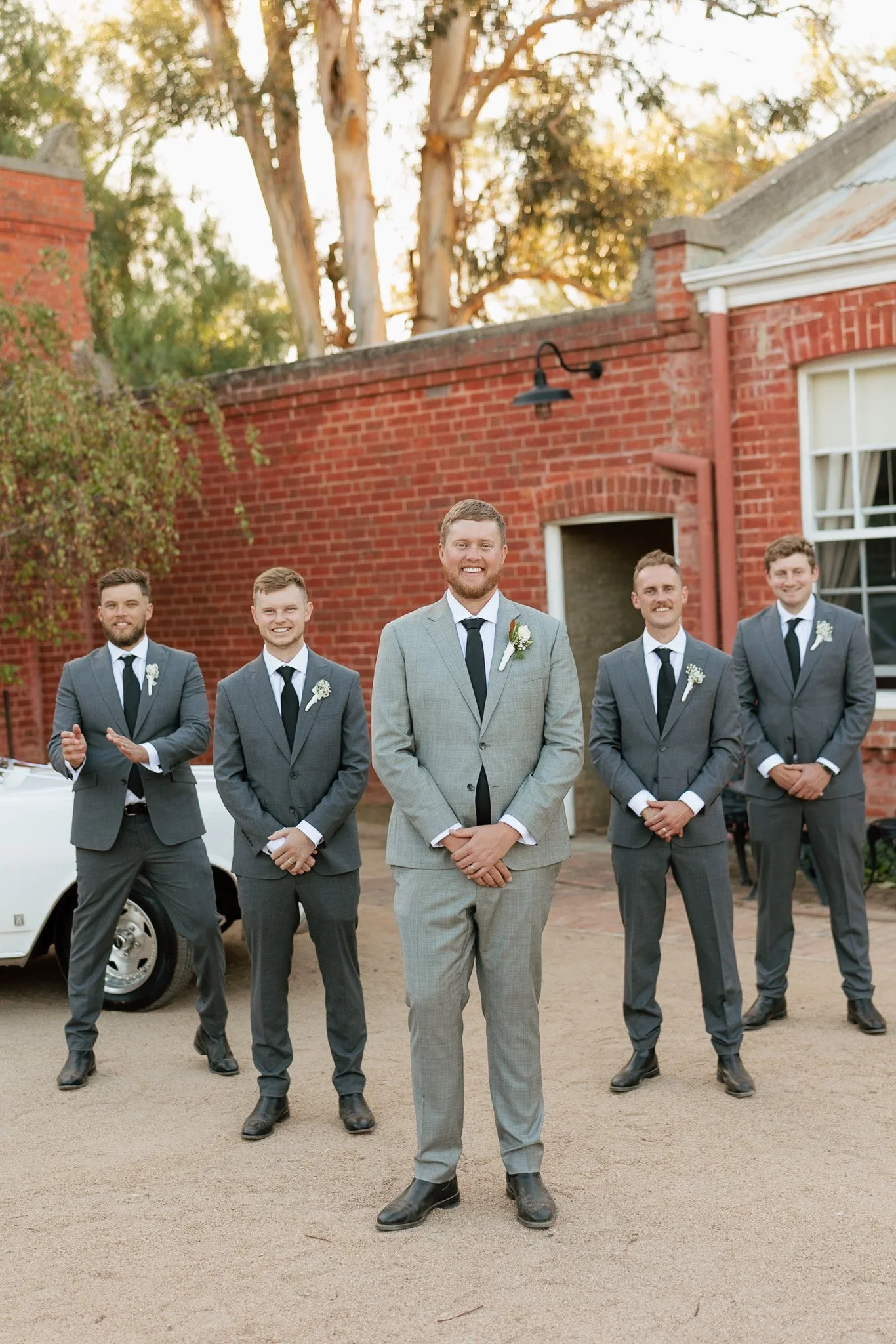 A groom and four groomsmen dressed in suits, standing outdoors in front of a brick building, with trees and sunlight in the background.