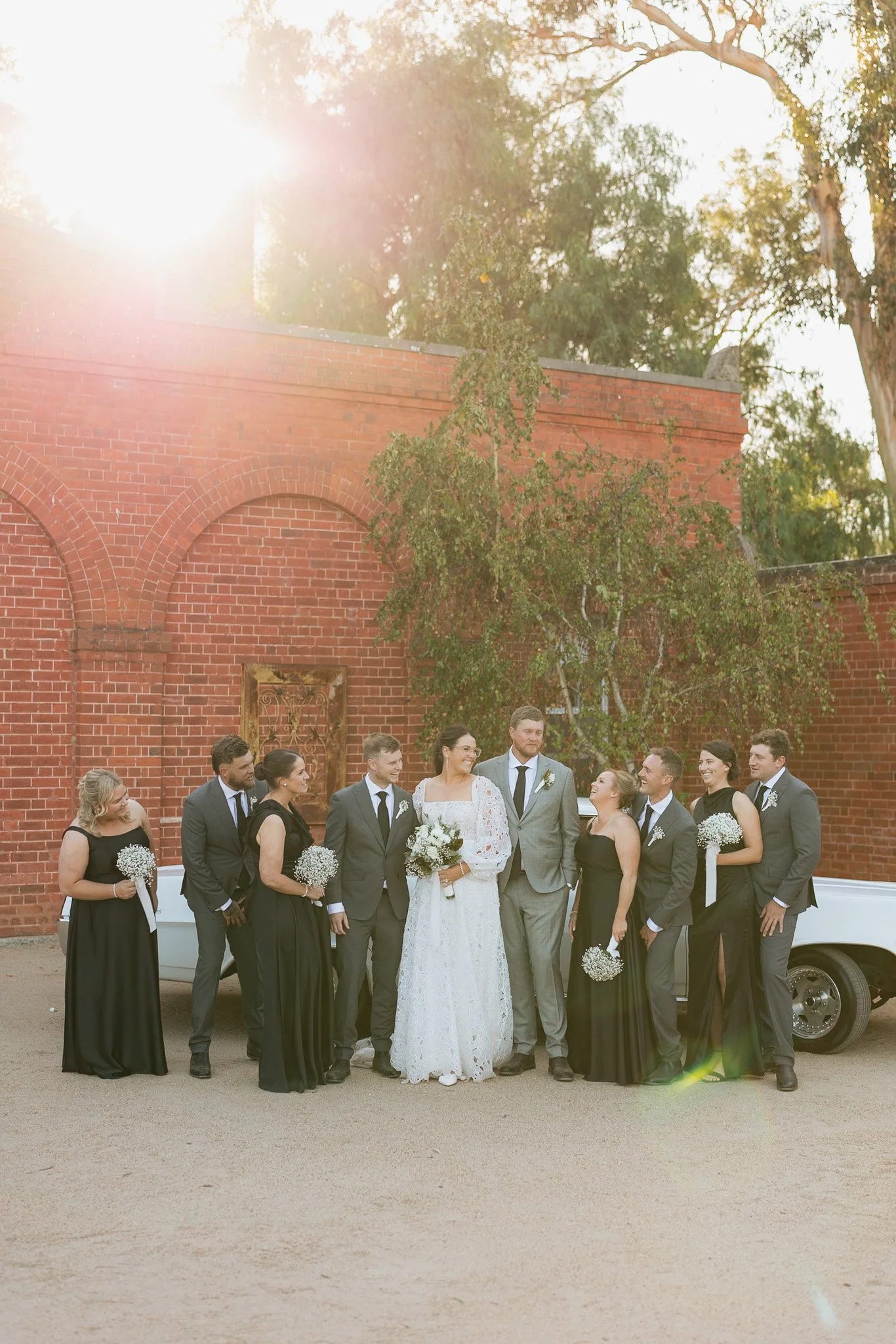 A group of wedding guests, including the bride and groom, standing outdoors in front of a red brick building with trees and sunlight in the background.