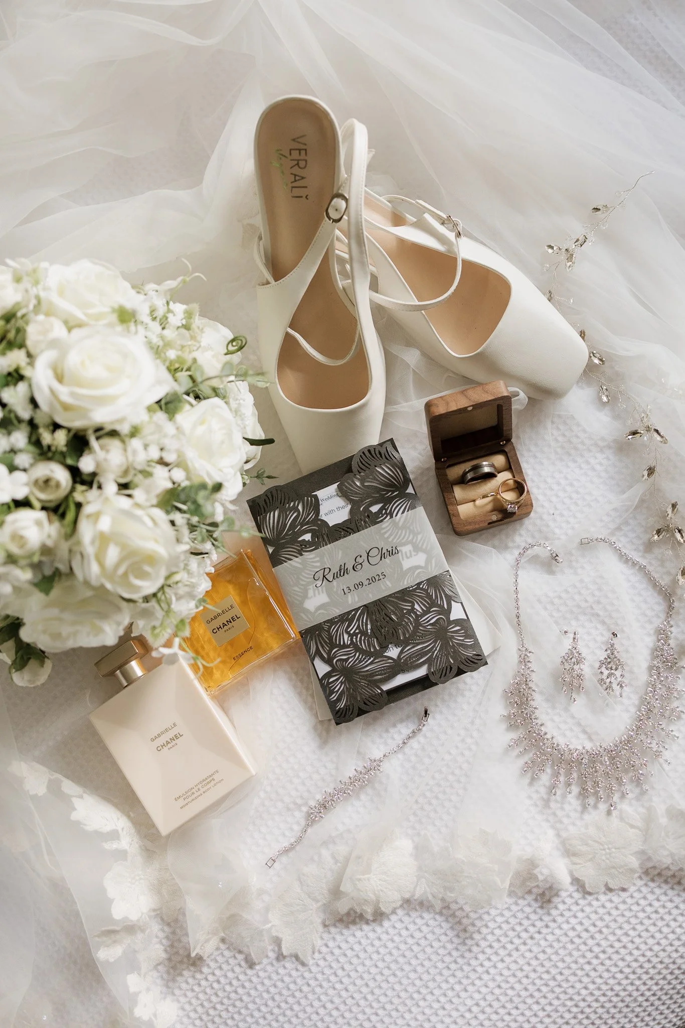 Wedding shoes, a bouquet of white roses, perfume, jewelry including earrings, a ring, and a bracelet, a wedding invitation and a small wooden box, all arranged on white fabric with lace and decorative elements.