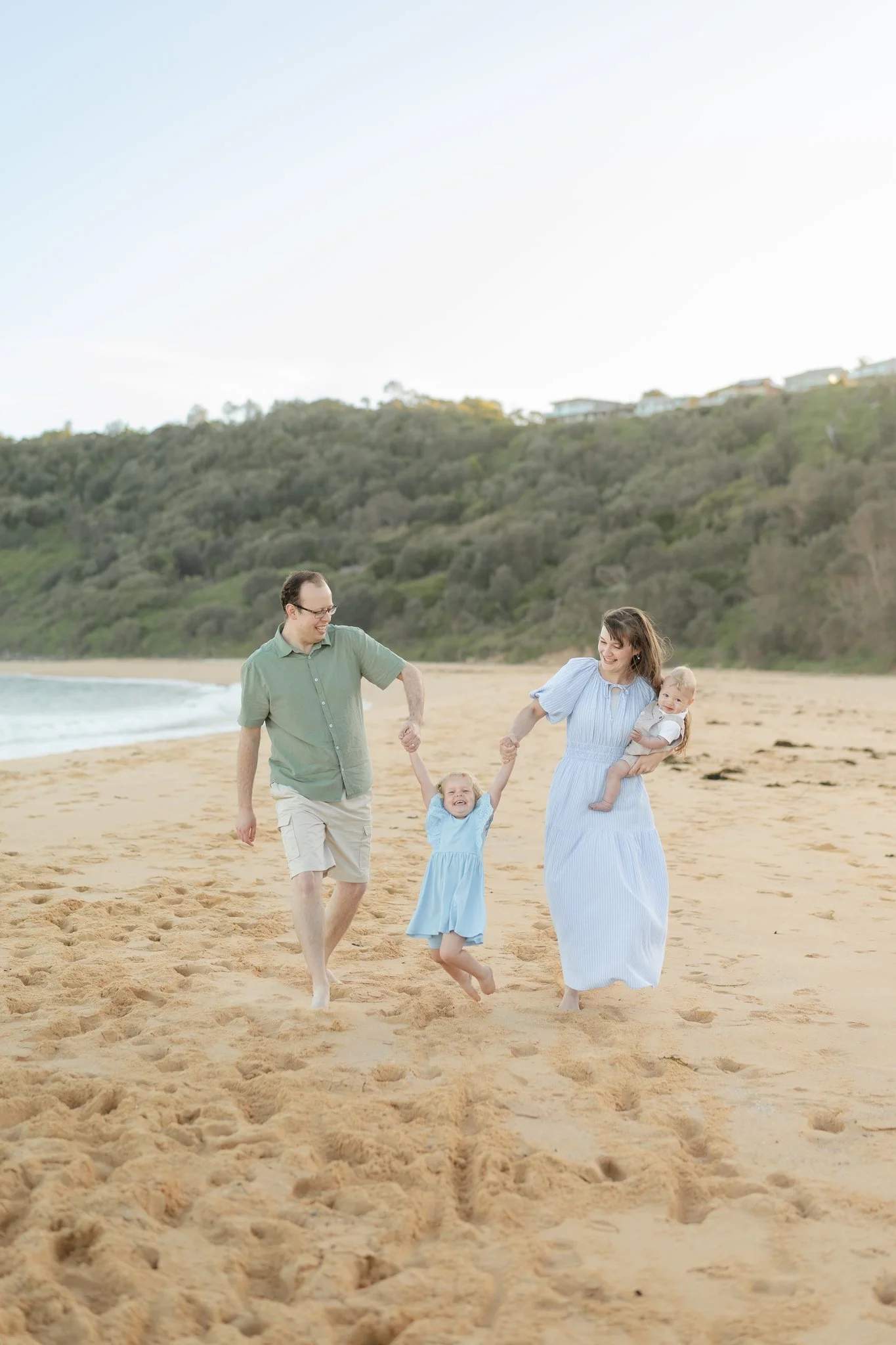 A family of four playing on the beach, with a man and woman holding their two young children by the hand, smiling and enjoying a sunny day.