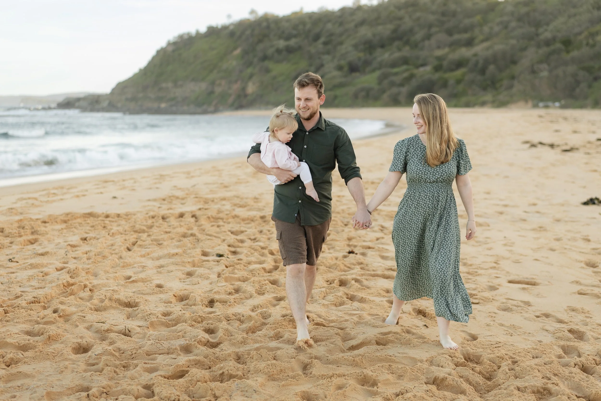 A family of three walking on a sandy beach, holding hands, with a man carrying a young girl in his arms, and a woman smiling beside them, in a scenic coastal area with hills and waves in the background.