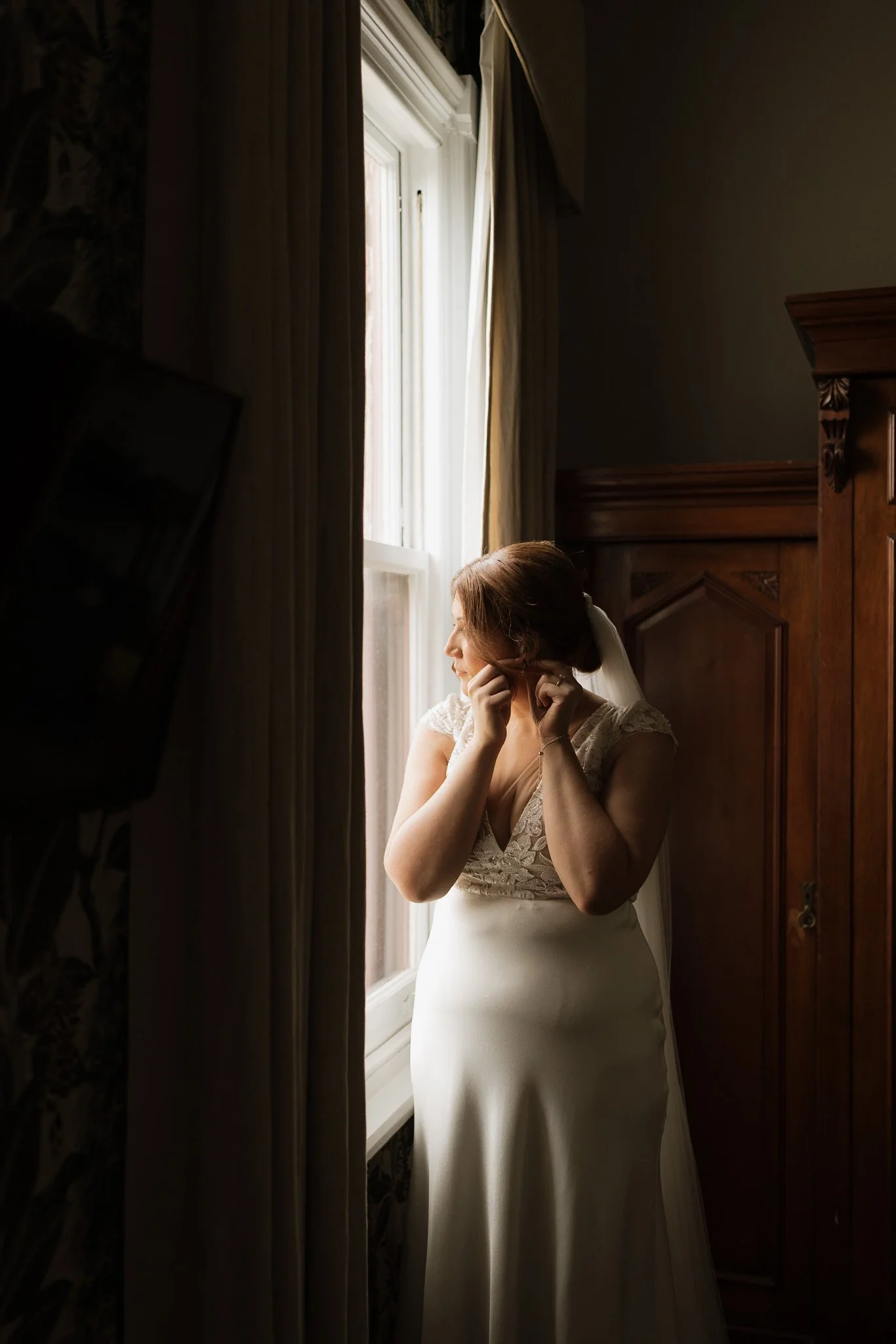 A bride in a white wedding dress looks out a window while touching her face gently.