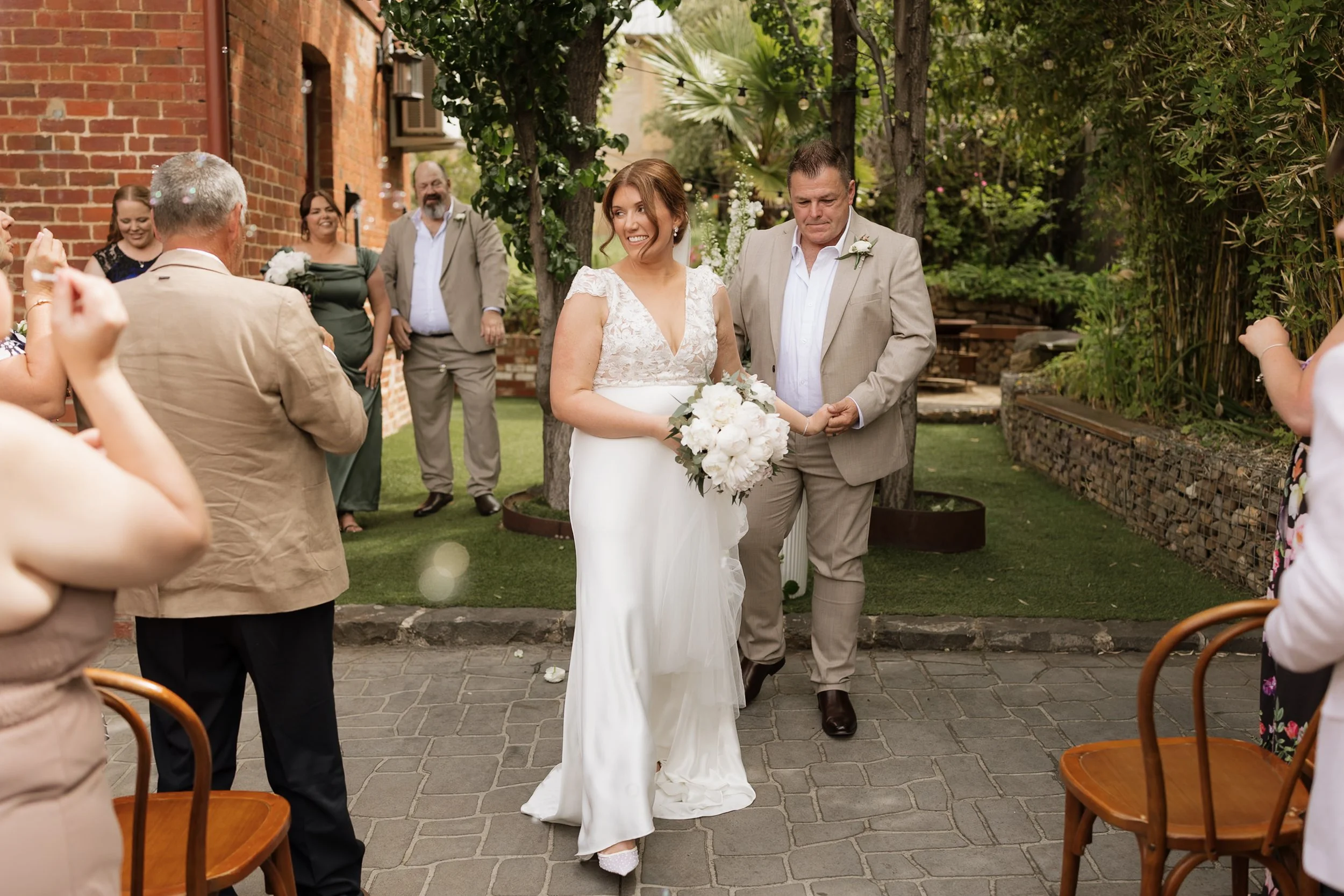 A bride in a white wedding dress holding a bouquet of white flowers walks down the aisle at an outdoor wedding ceremony, accompanied by a man in a light gray suit.