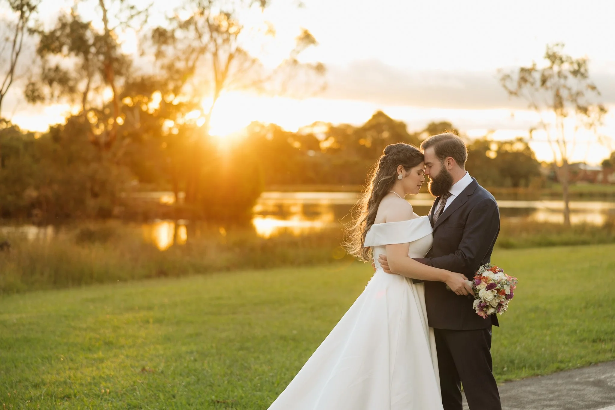 A bride and groom standing close together outdoors during sunset, with trees and a lake in the background. The bride is holding a bouquet of flowers, and both are dressed in wedding attire with their foreheads touching.