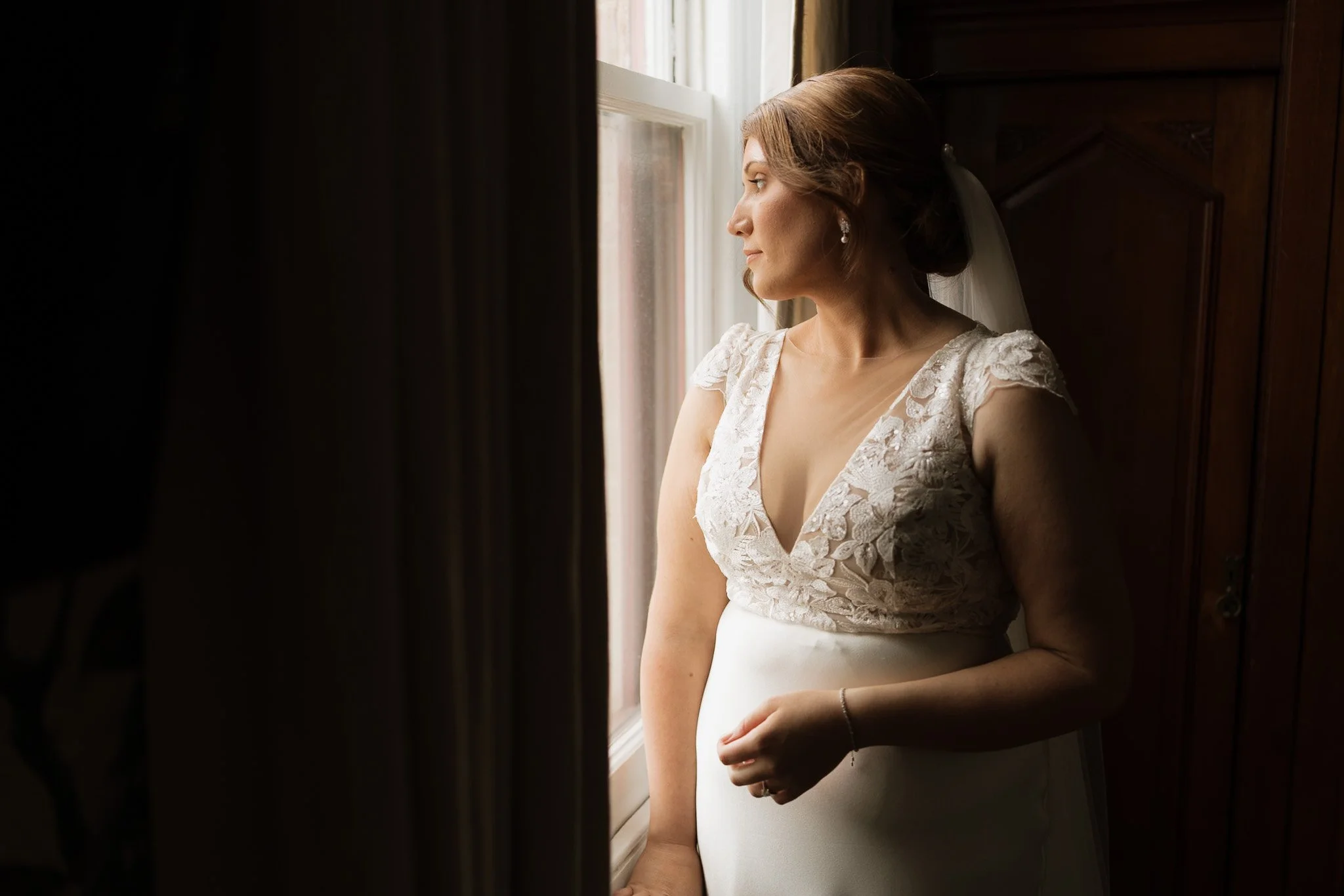 A bride in a lace wedding dress standing by a window, looking outside, with natural light illuminating her face and side profile.