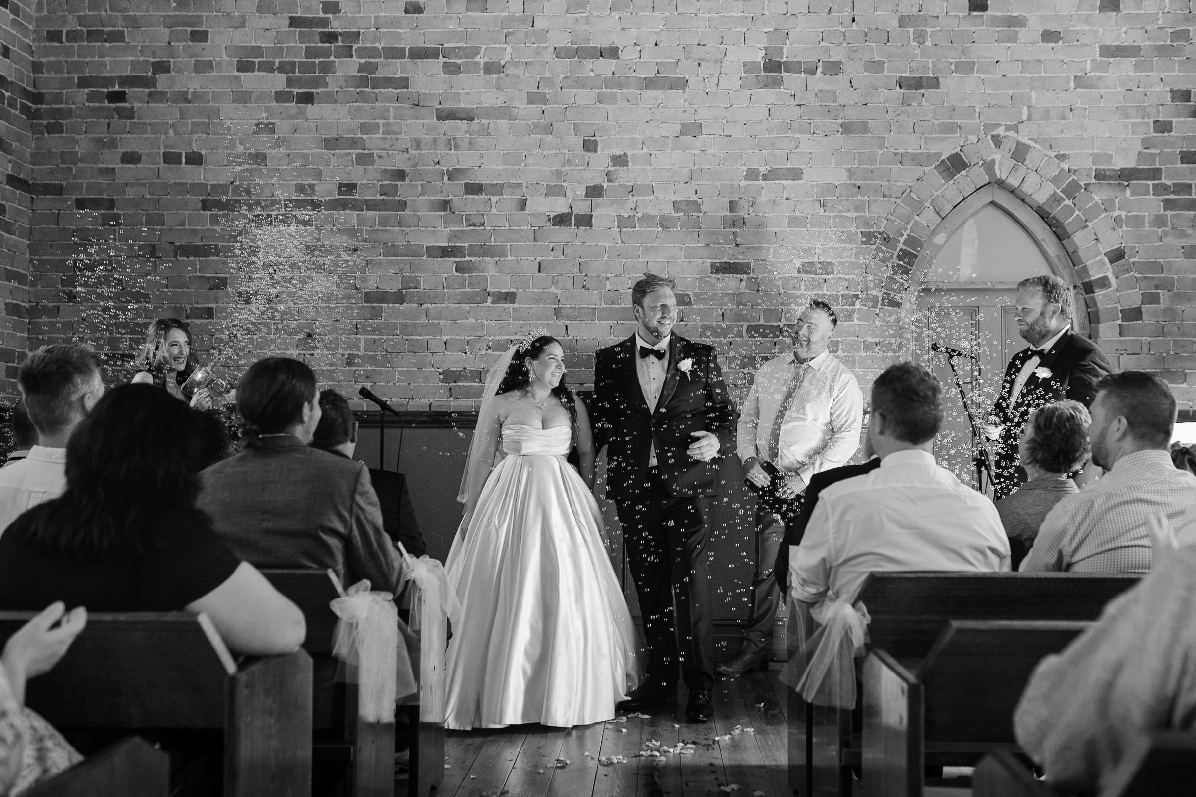 A wedding ceremony in a church with brick walls, a bride and groom exchanging vows, surrounded by friends and family, with confetti falling.
