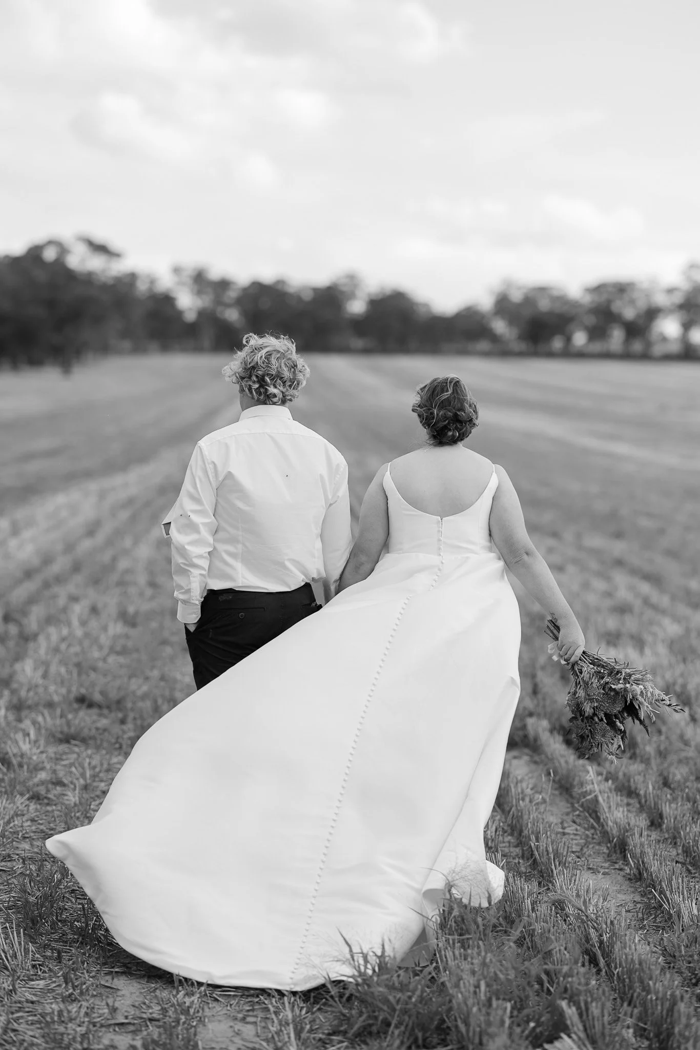 A black and white photo of a bride and groom walking away together across a grassy field. The bride wears a long wedding dress and holds a bouquet of flowers. The groom wears a white shirt and dark pants. Trees are visible in the background, and the 