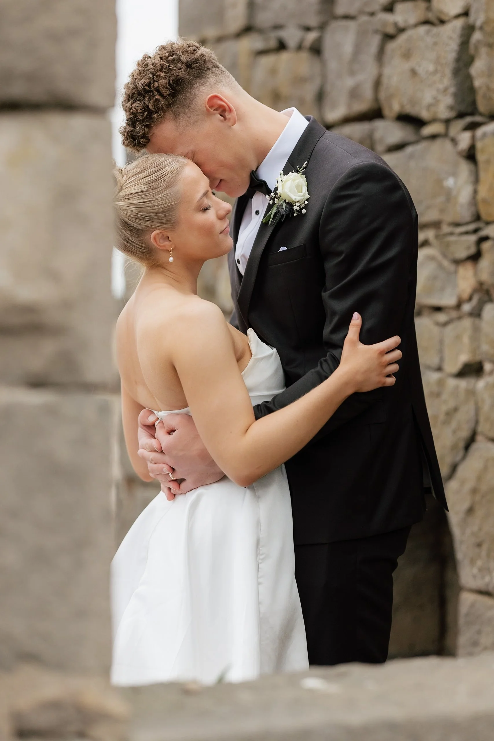 A bride and groom sharing a romantic moment outdoors, embracing each other with their foreheads touching, near a stone wall.