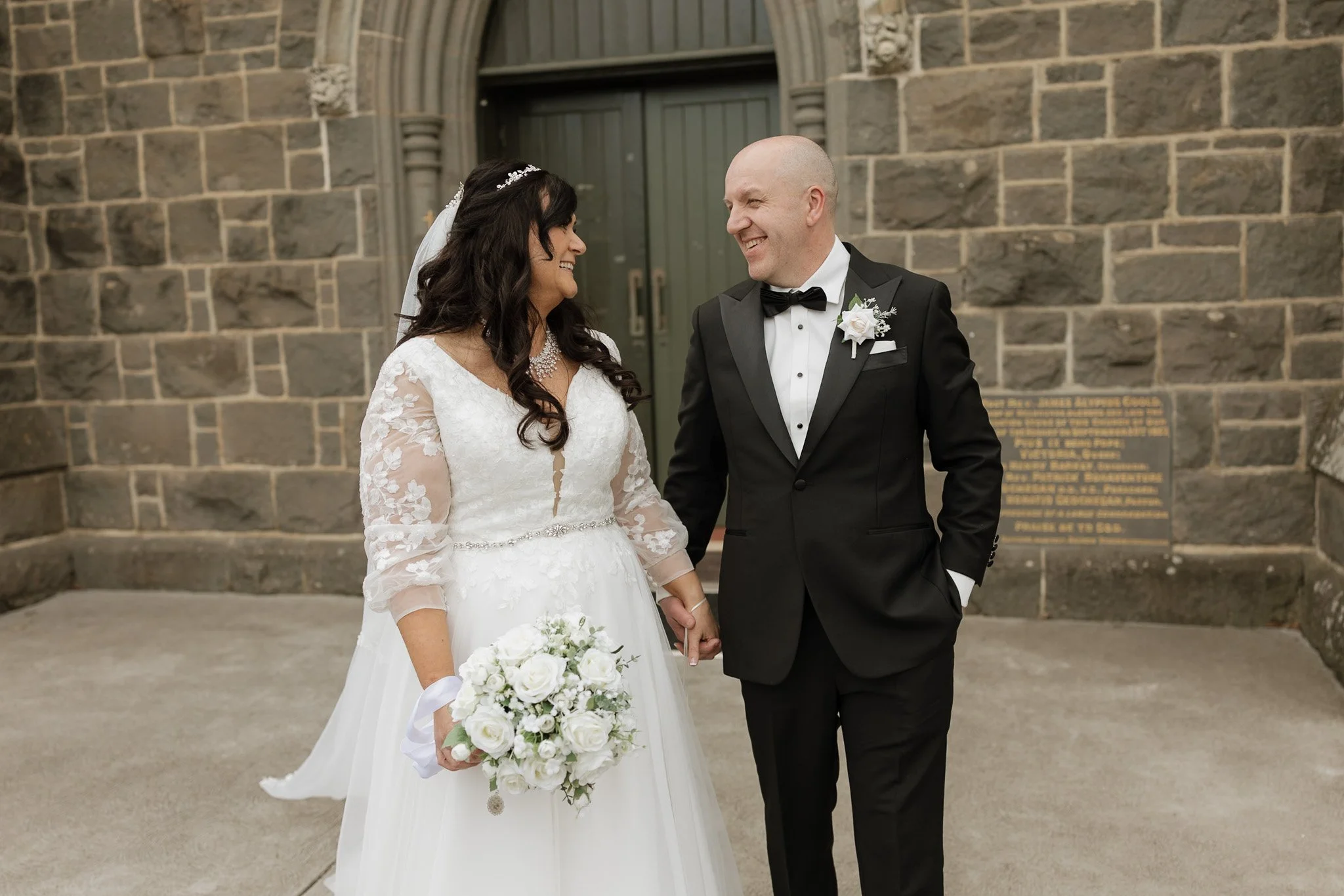A bride and groom smiling and holding hands outside a stone building, dressed in wedding attire with the bride holding a bouquet of white roses.