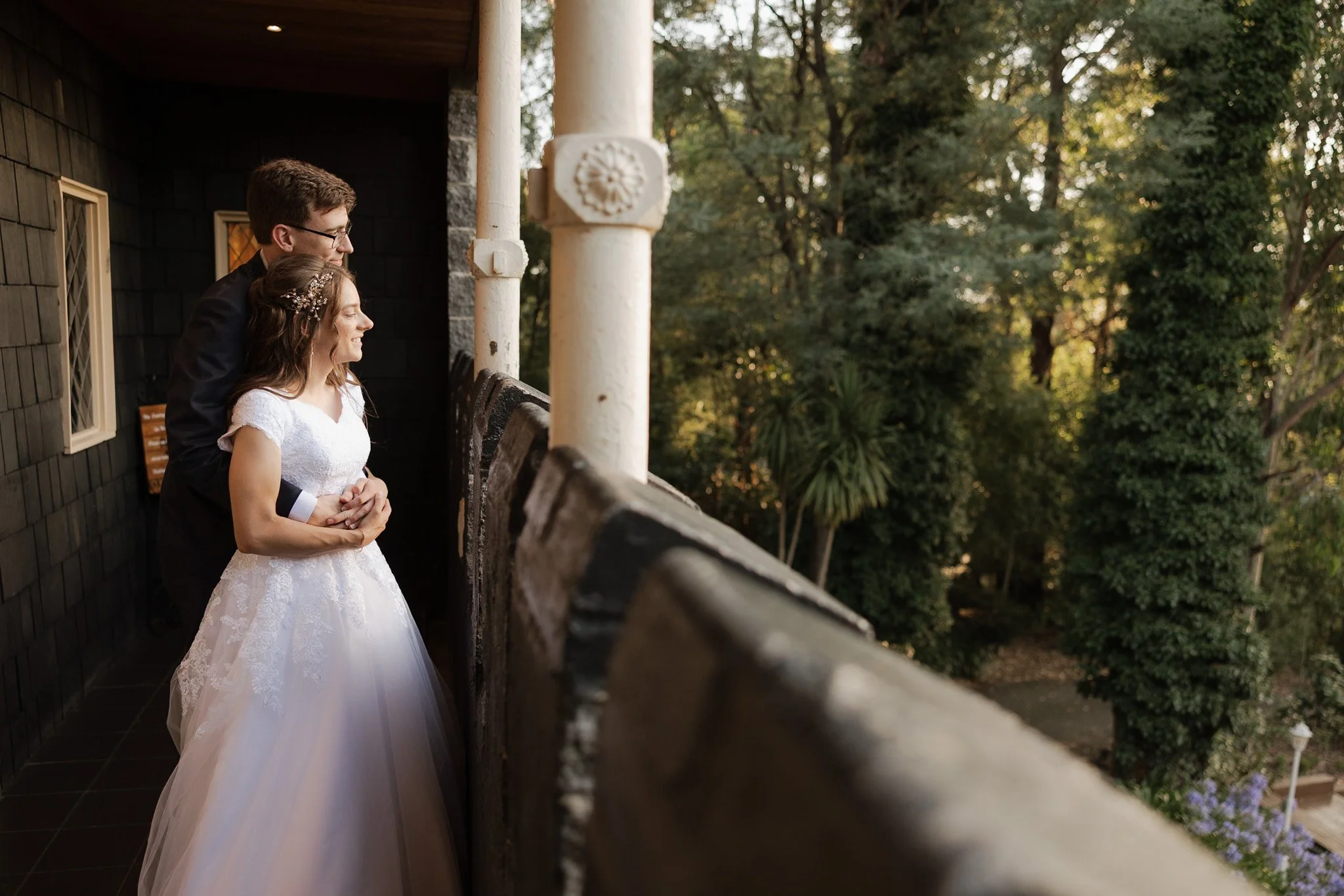 A bride in a white wedding dress and a man in a black suit standing on a balcony with greenery in the background at sunset.