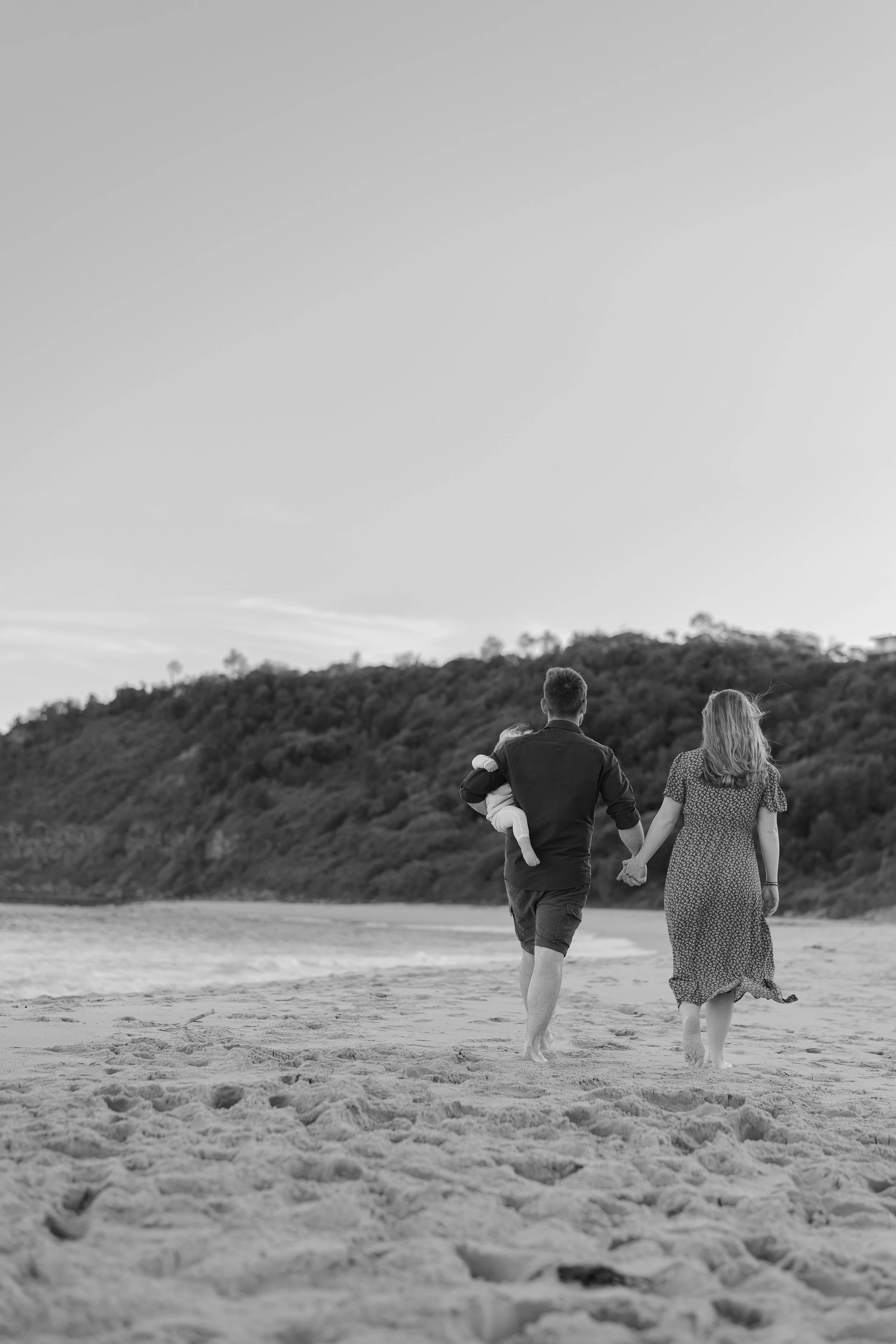 A black and white photograph of a family walking on a sandy beach during sunset, with a man holding a child and a woman holding the man's hand.