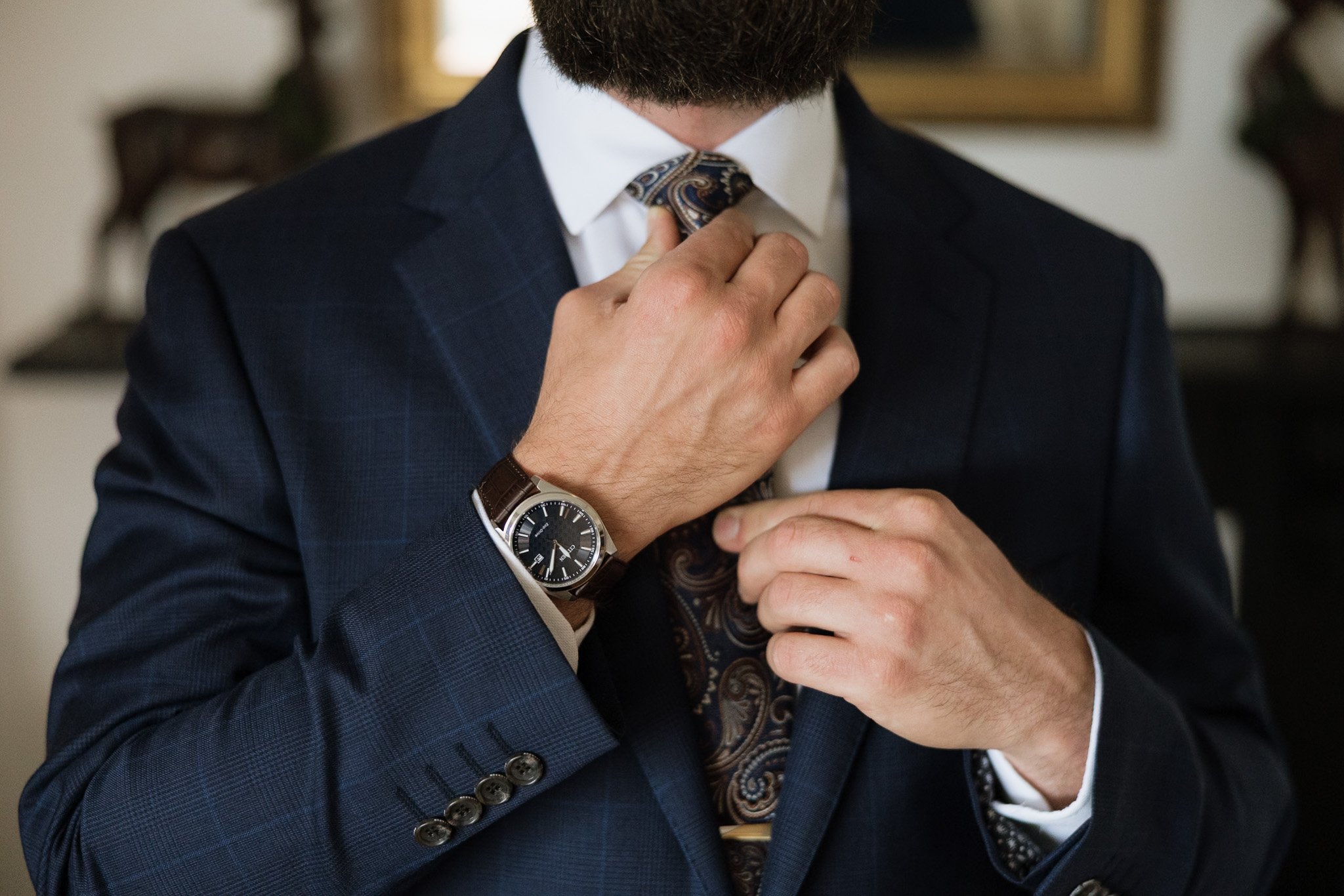 Close-up of a man adjusting his paisley tie while wearing a dark blue suit jacket and a white shirt, with a large wristwatch on his left wrist.