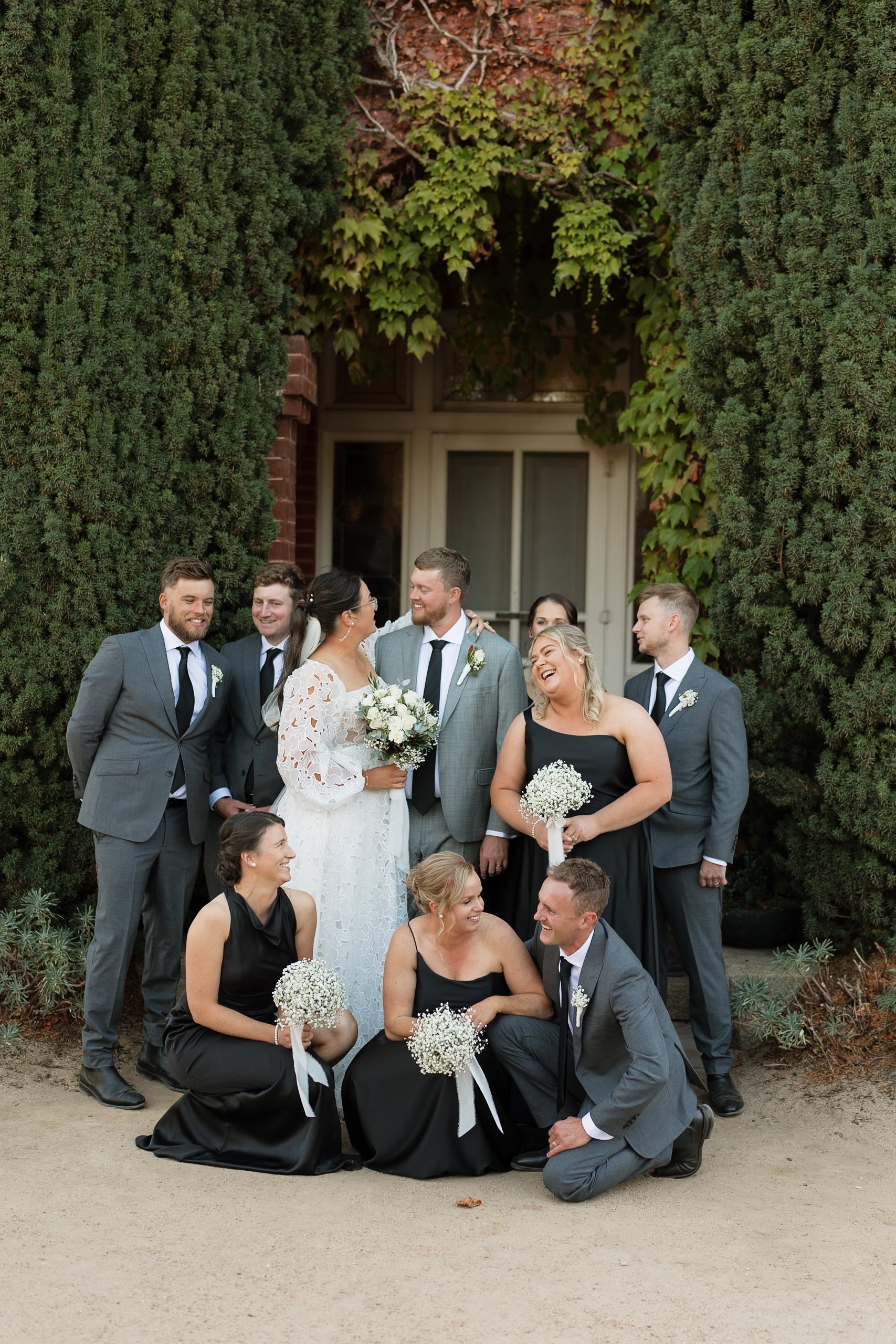 A wedding party poses outdoors in front of green hedges and an arch covered with ivy. The bride and groom are at the center, smiling and looking at each other, surrounded by bridesmaids and groomsmen.