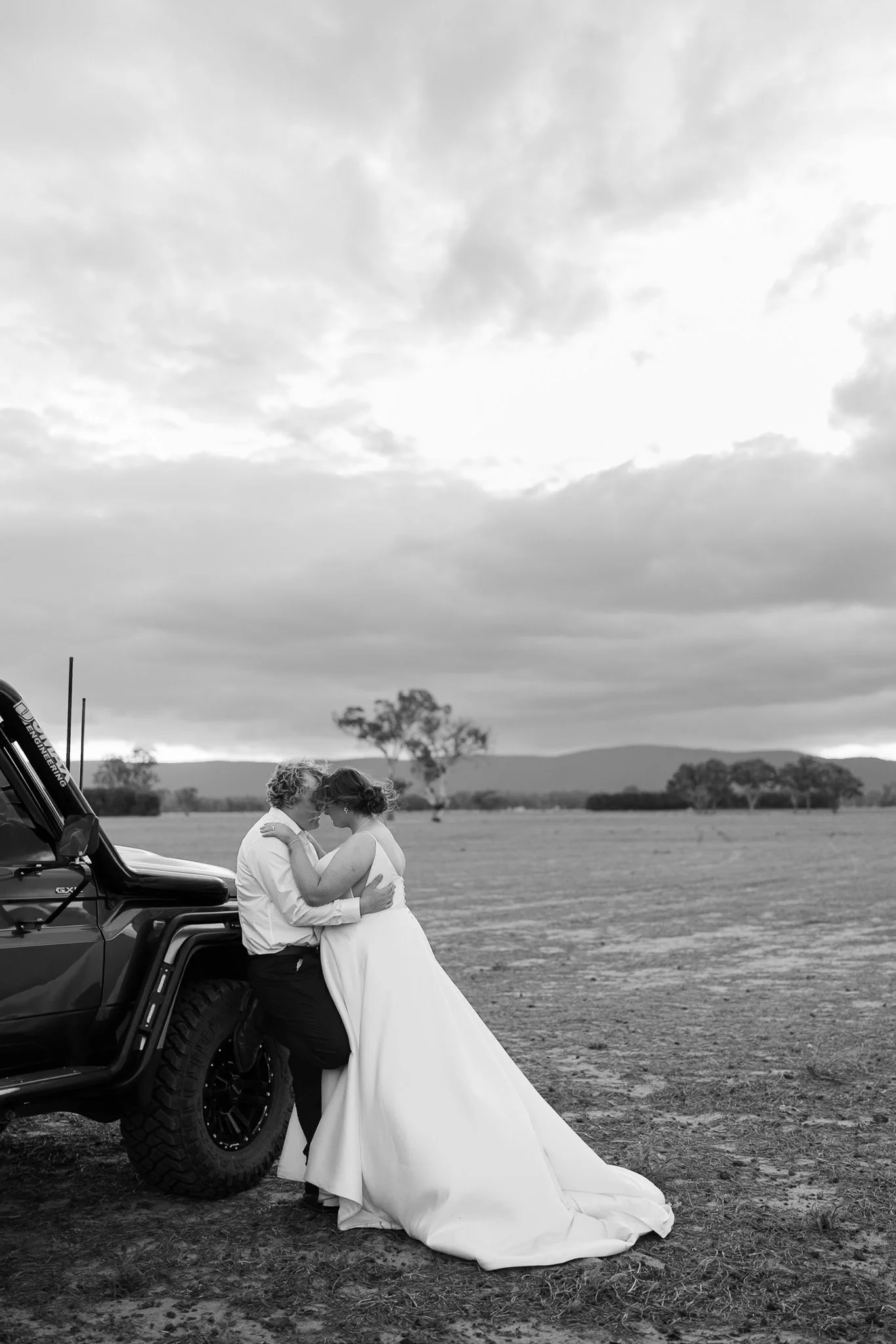 A black-and-white photo of a bride and groom embracing outdoors next to a vehicle, with a cloudy sky and distant mountains in the background.