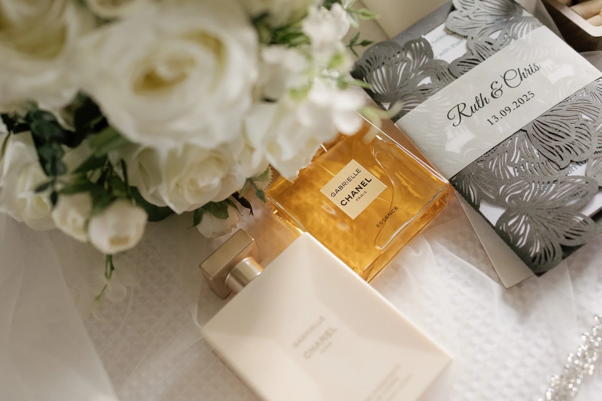 A bouquet of white flowers, a bottle of Chanel Gabrielle perfume, and a wedding invitation with the names Ruth and Chris, and the date 13.09.2025.