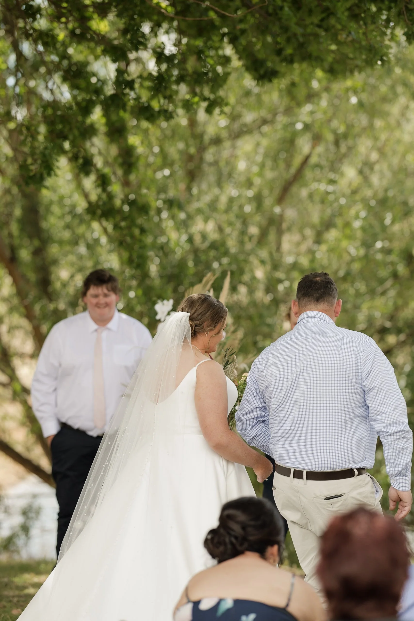 A wedding ceremony outdoors with a bride and groom holding hands and standing under a canopy of tree leaves, surrounded by guests.