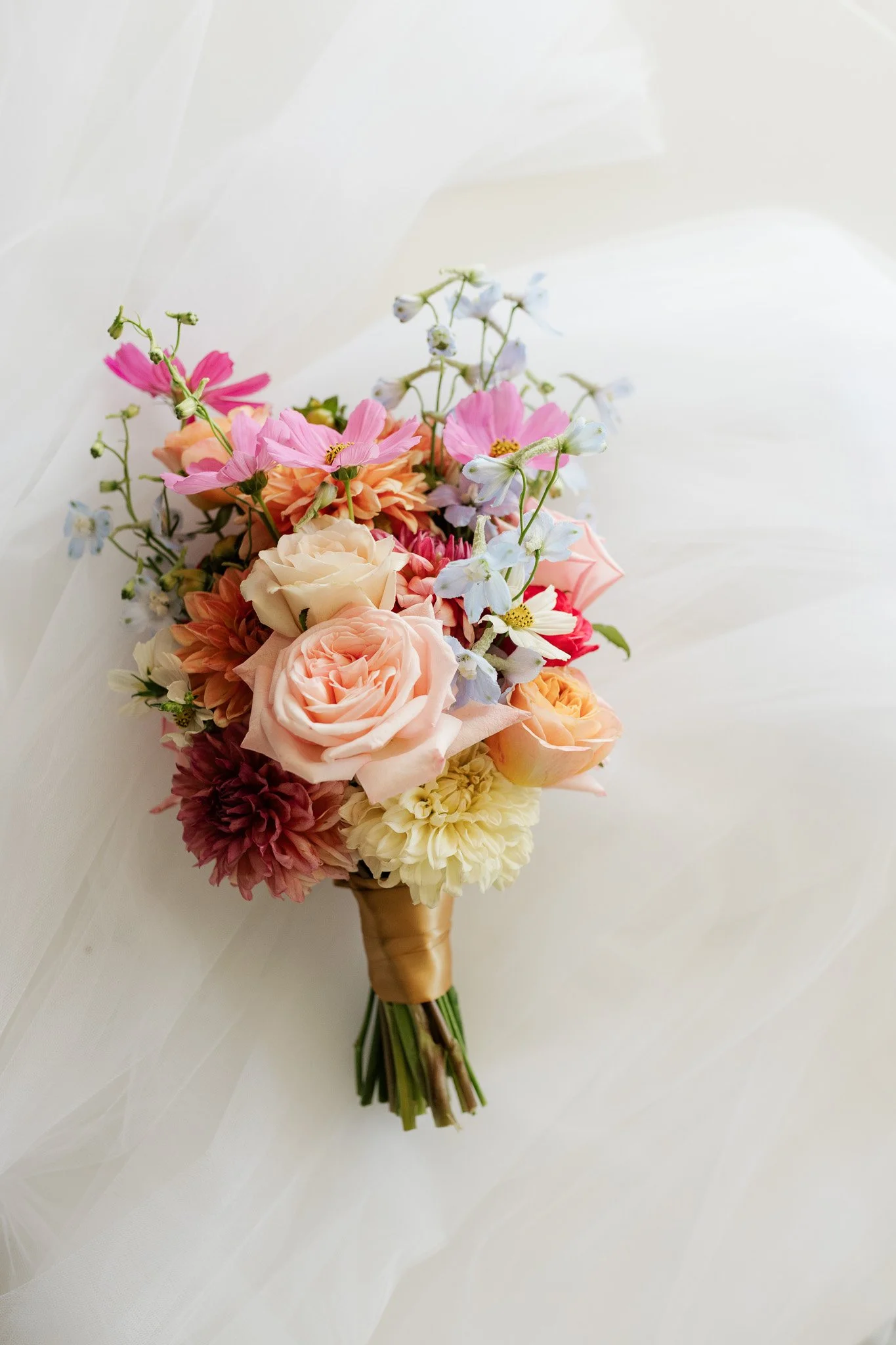 A bridal bouquet with pink, peach, ivory, and cream flowers, wrapped with a satin ribbon, displayed on a white surface.