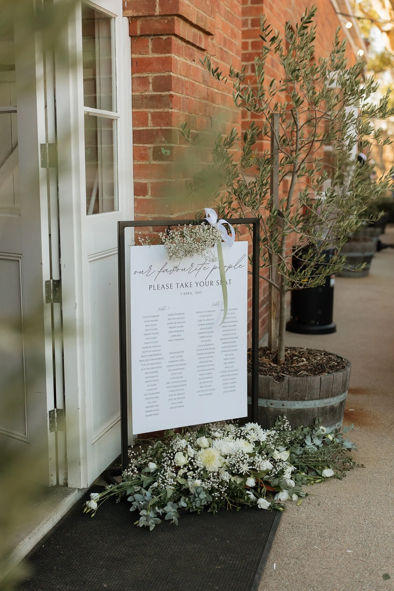 A wedding seating chart sign in front of a brick building entrance, decorated with white flowers and greenery at the base, with a small bouquet attached to the top of the sign and tied with a white ribbon.