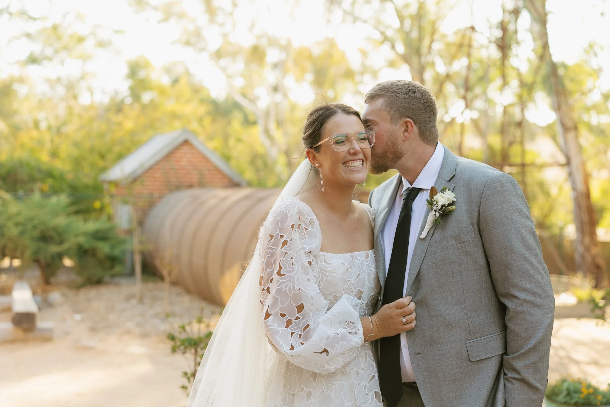 A bride and groom sharing a kiss outdoors, with the groom gently kissing the bride's temple. The bride is wearing glasses, a white lace wedding dress, and a veil, while the groom is dressed in a gray suit with a white shirt and a black tie. The backg