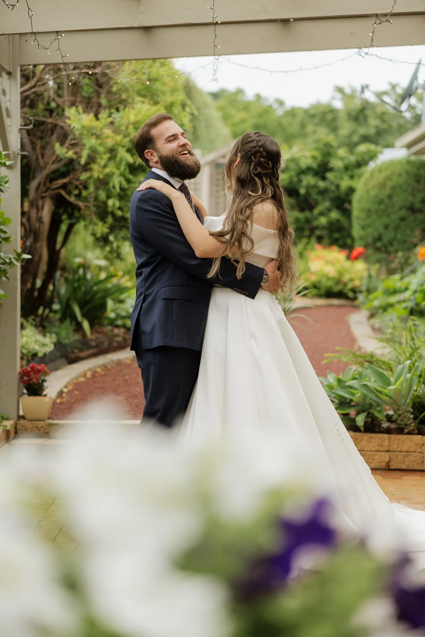 A couple dancing outdoors, smiling at each other during their wedding, with greenery and string lights in the background.