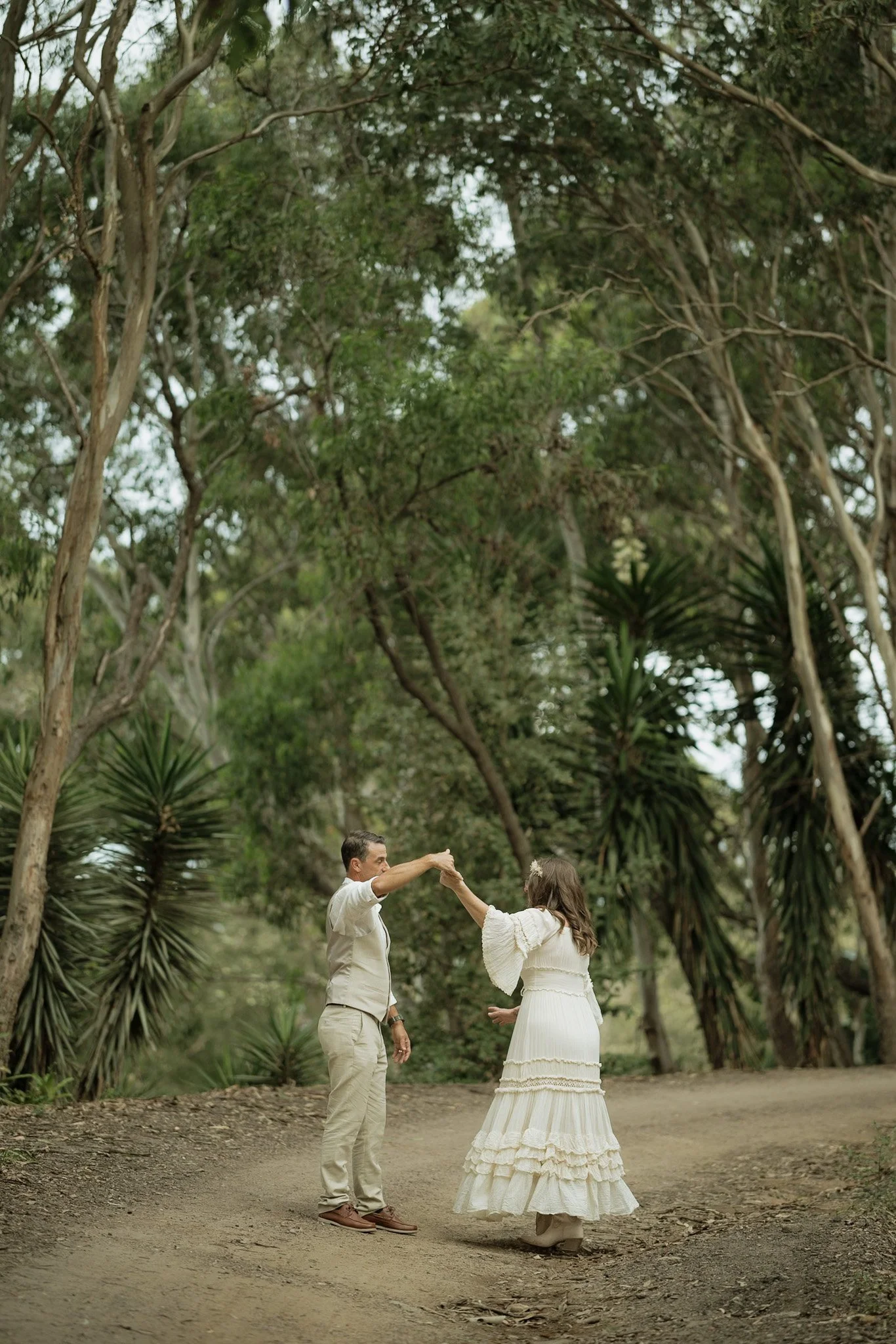 A couple dancing outdoors in a lush, wooded area. The man is in beige clothing and the woman is wearing a white dress. They are holding hands and spinning together on a dirt path surrounded by green trees and plants.