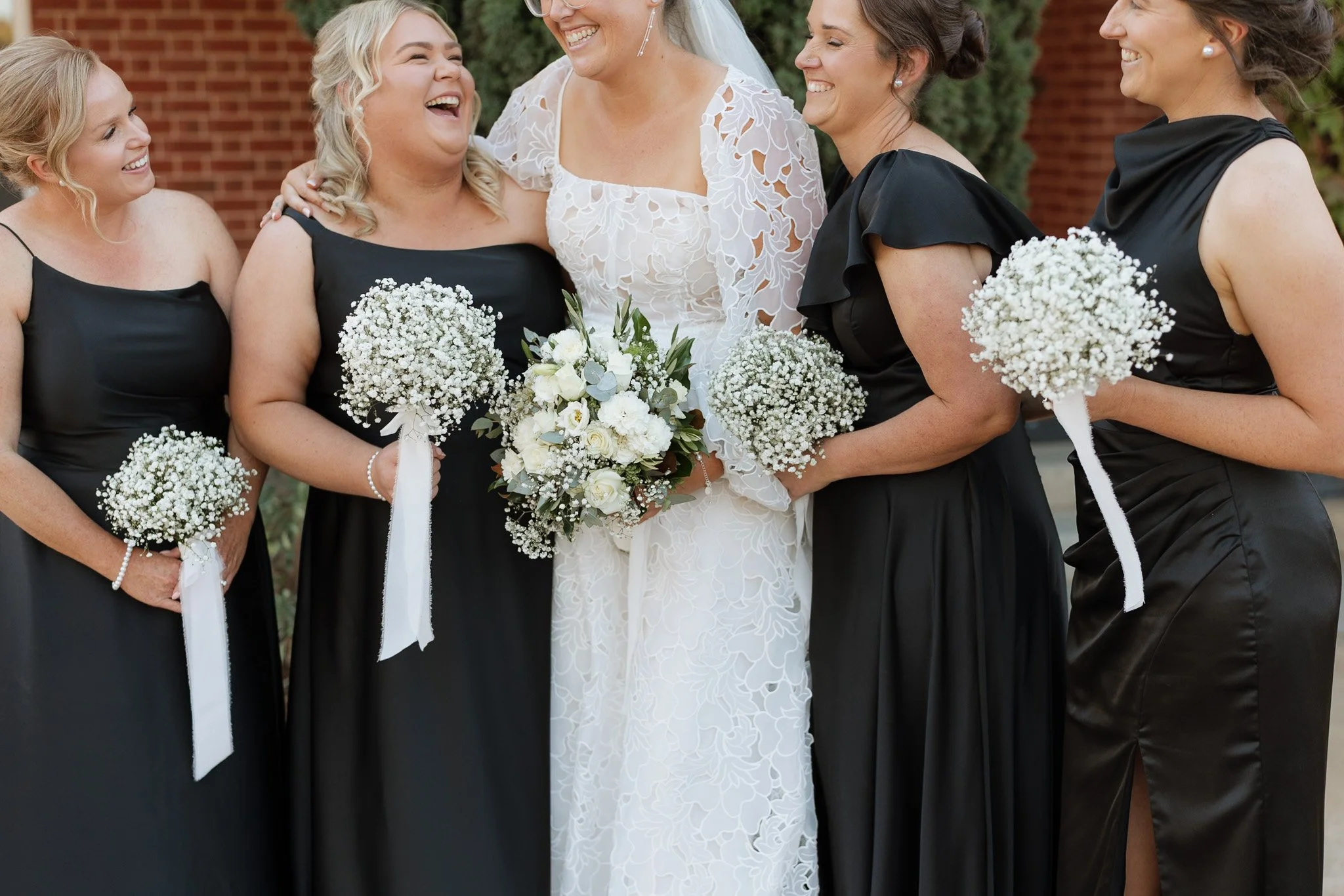 A bride in a white wedding dress smiling while standing among four women in black dresses, all holding small bouquets of white flowers, outdoors in front of a brick wall.