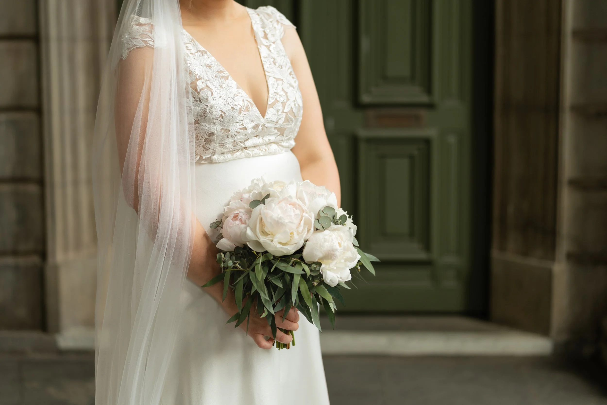 A bride in a white wedding dress holding a bouquet of white peonies and eucalyptus leaves, standing outside near a green door.