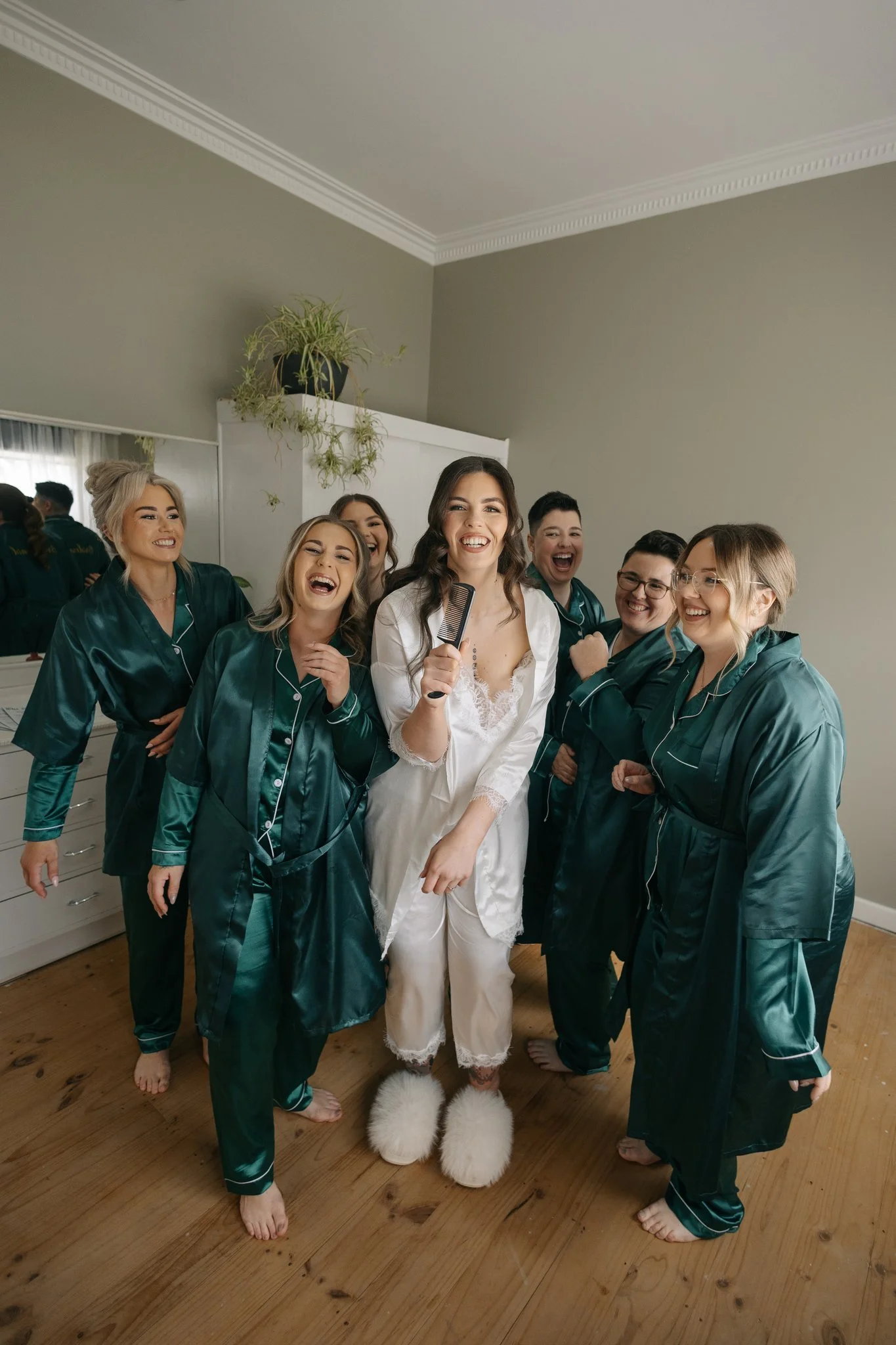 A bride in white pajamas and fluffy slippers holding a hairbrush, surrounded by six bridesmaids in teal pajamas, all smiling and posing on a wooden floor inside a home.
