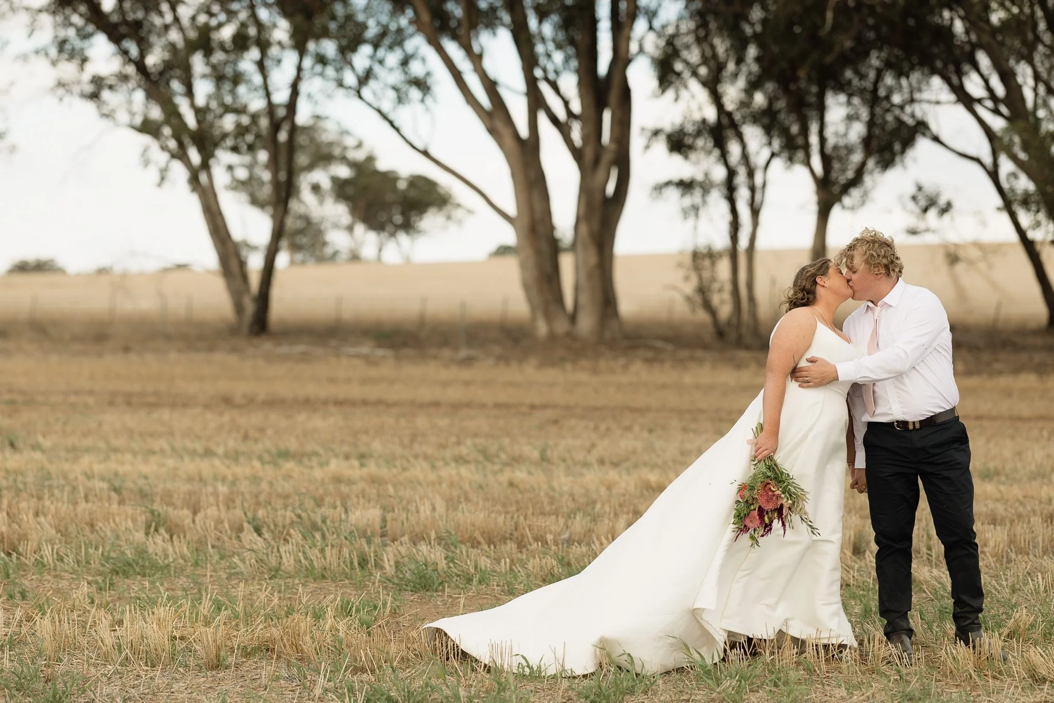 A bride and groom share a kiss outdoors in a field surrounded by trees, with the bride holding a bouquet of flowers.