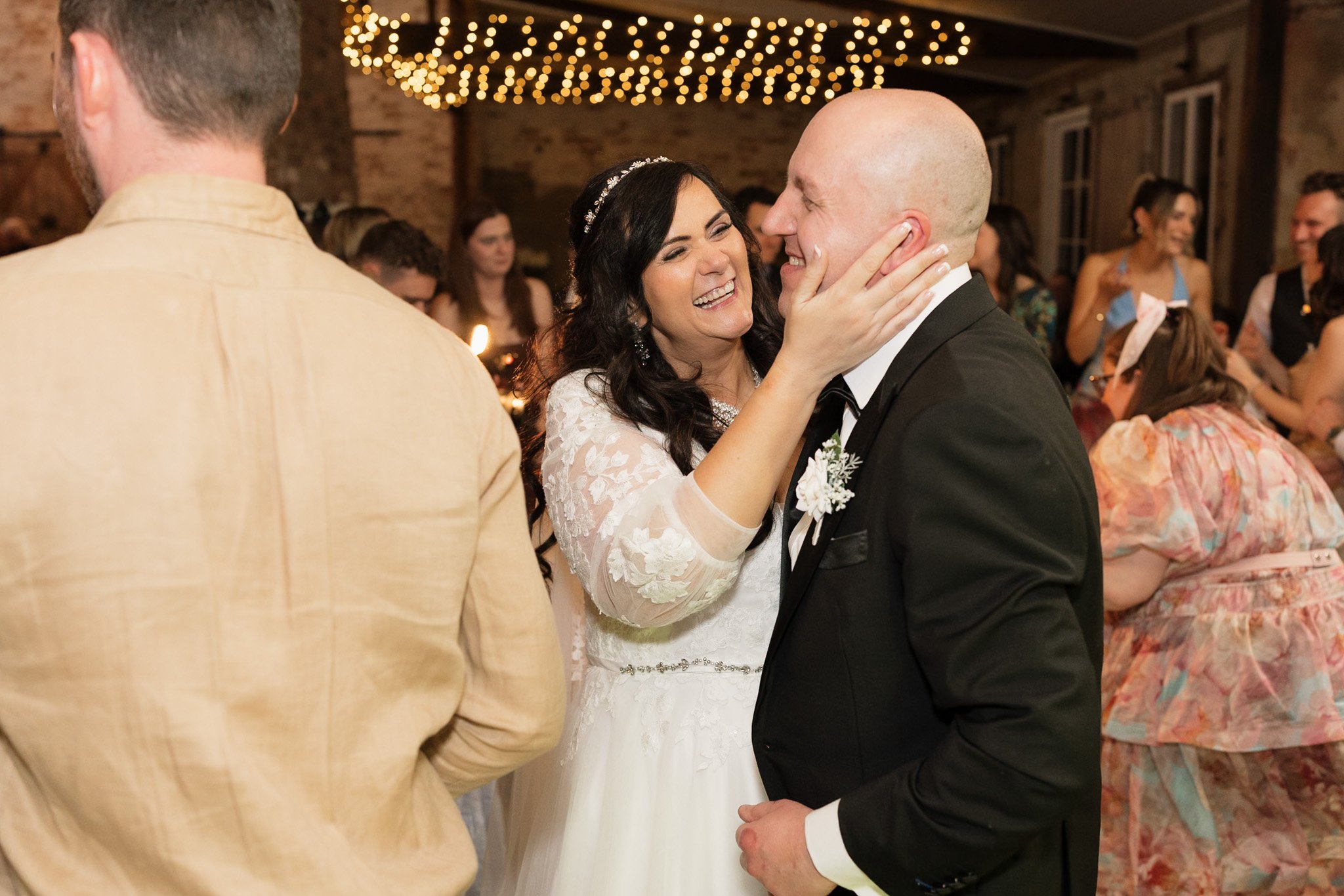 Bride and groom smiling and dancing at their wedding reception, with guests in the background under string lights.