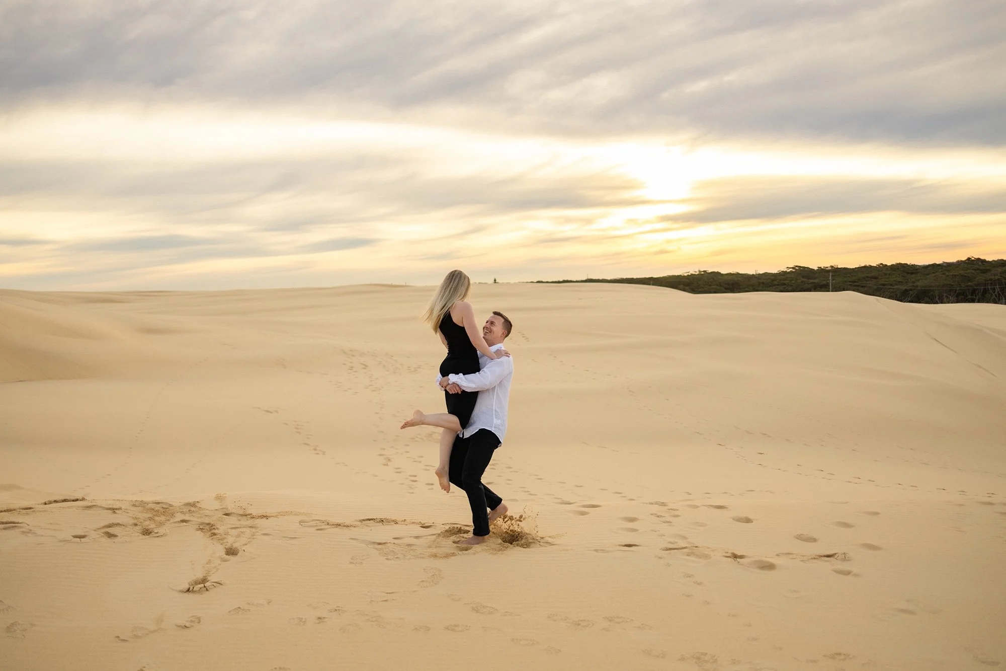 A couple is outdoors in a desert or sand dune landscape during sunset; the man is lifting the woman in his arms as they smile, with the woman in a black dress and the man in a white shirt and black pants, with footprints in the sand leading towards them.