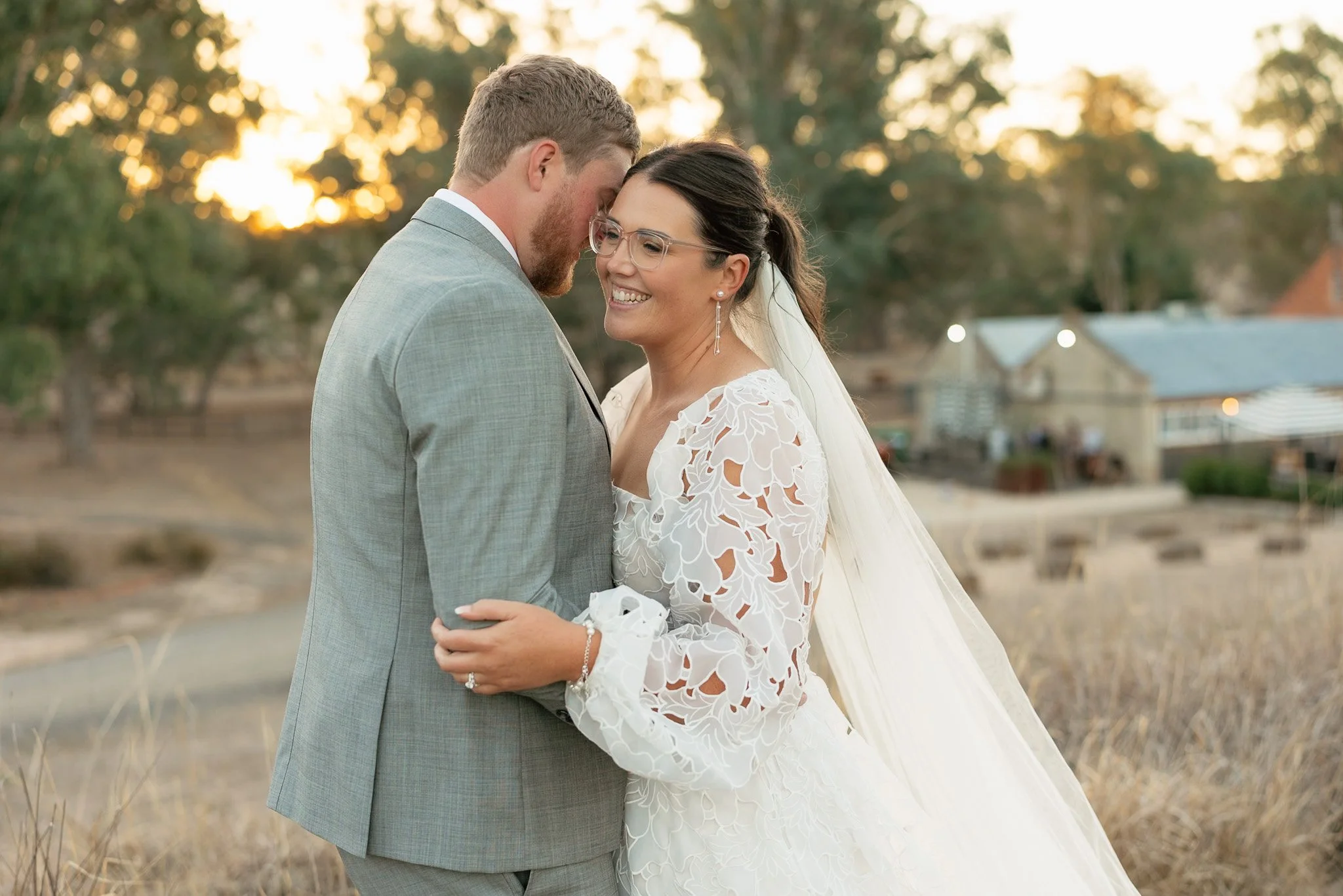 A bride and groom sharing a tender moment outdoors during sunset, with trees and a historic building in the background.