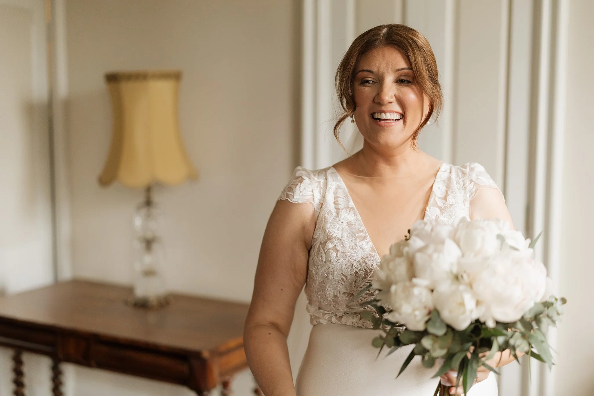 A woman in a wedding dress holding a bouquet of white flowers and greenery, standing indoors in front of a wooden stair railing and a beige wall with a lamp.