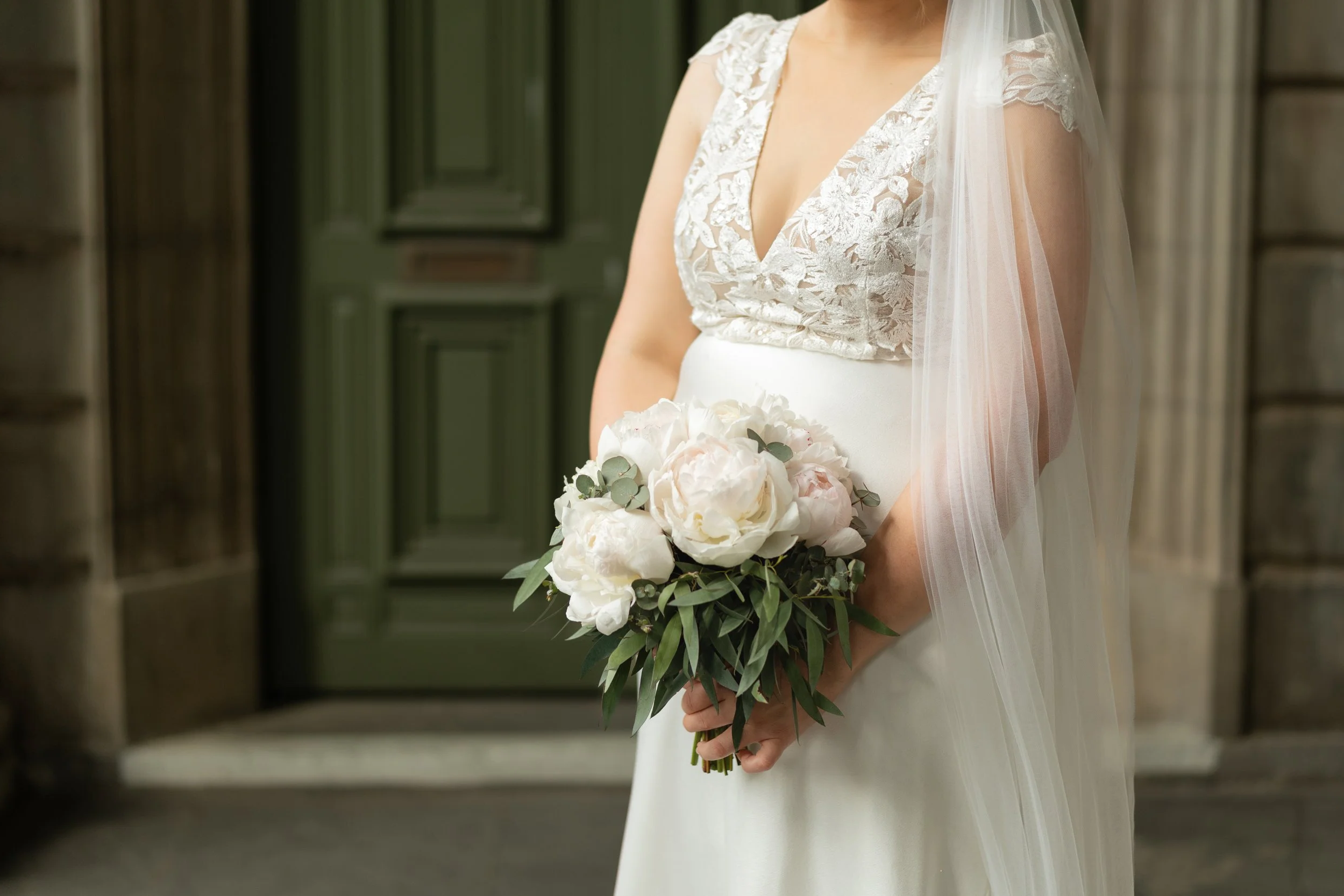 A bride in a white lace wedding dress holding a bouquet of white flowers and greenery inside a building with green and wooden paneling.