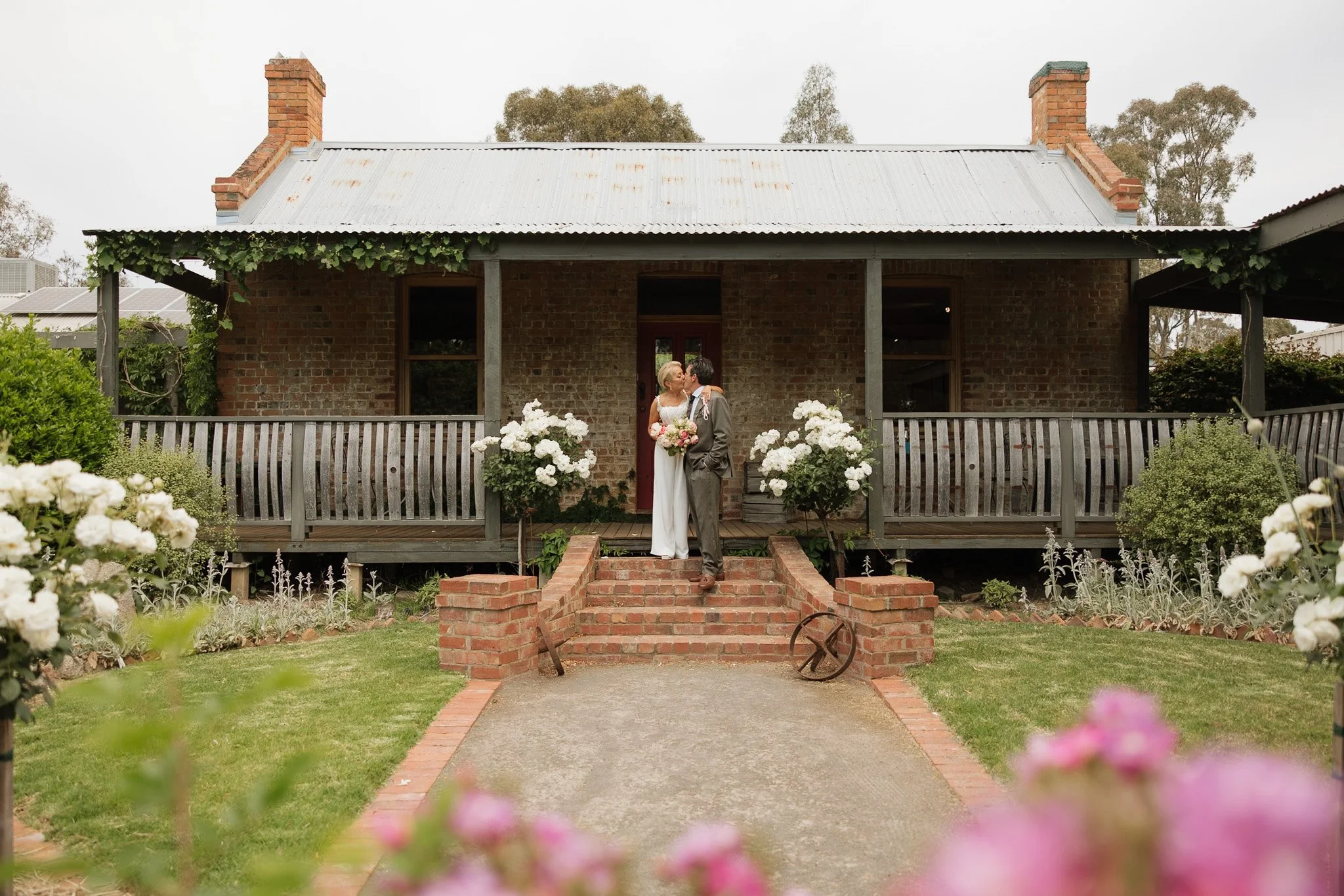 A bride and groom sharing a kiss on the porch of a brick house decorated with white flowers and greenery, with trees and other buildings in the background.