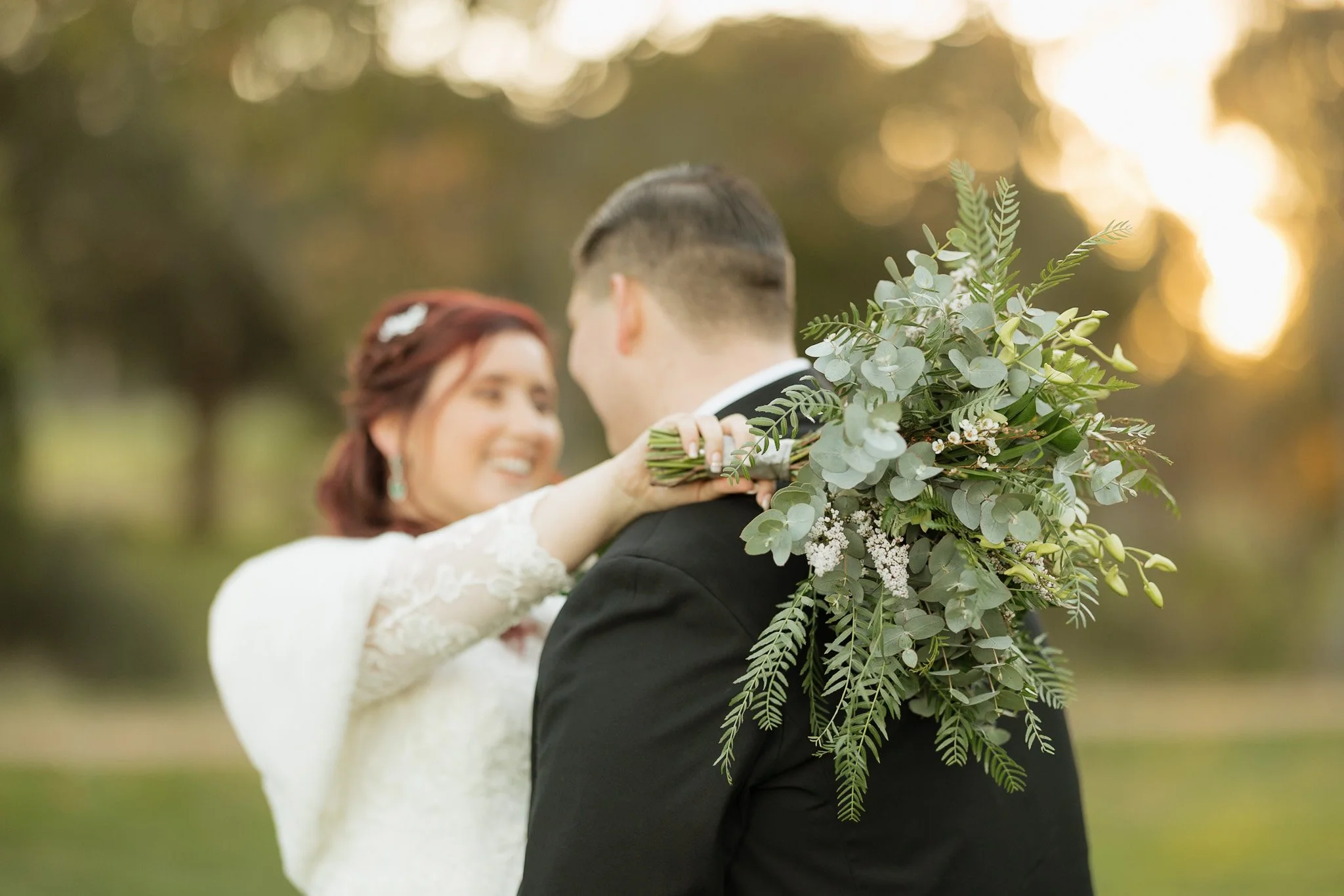 A bride and groom embracing outdoors during sunset, with the bride holding a large bouquet of greenery and white flowers.