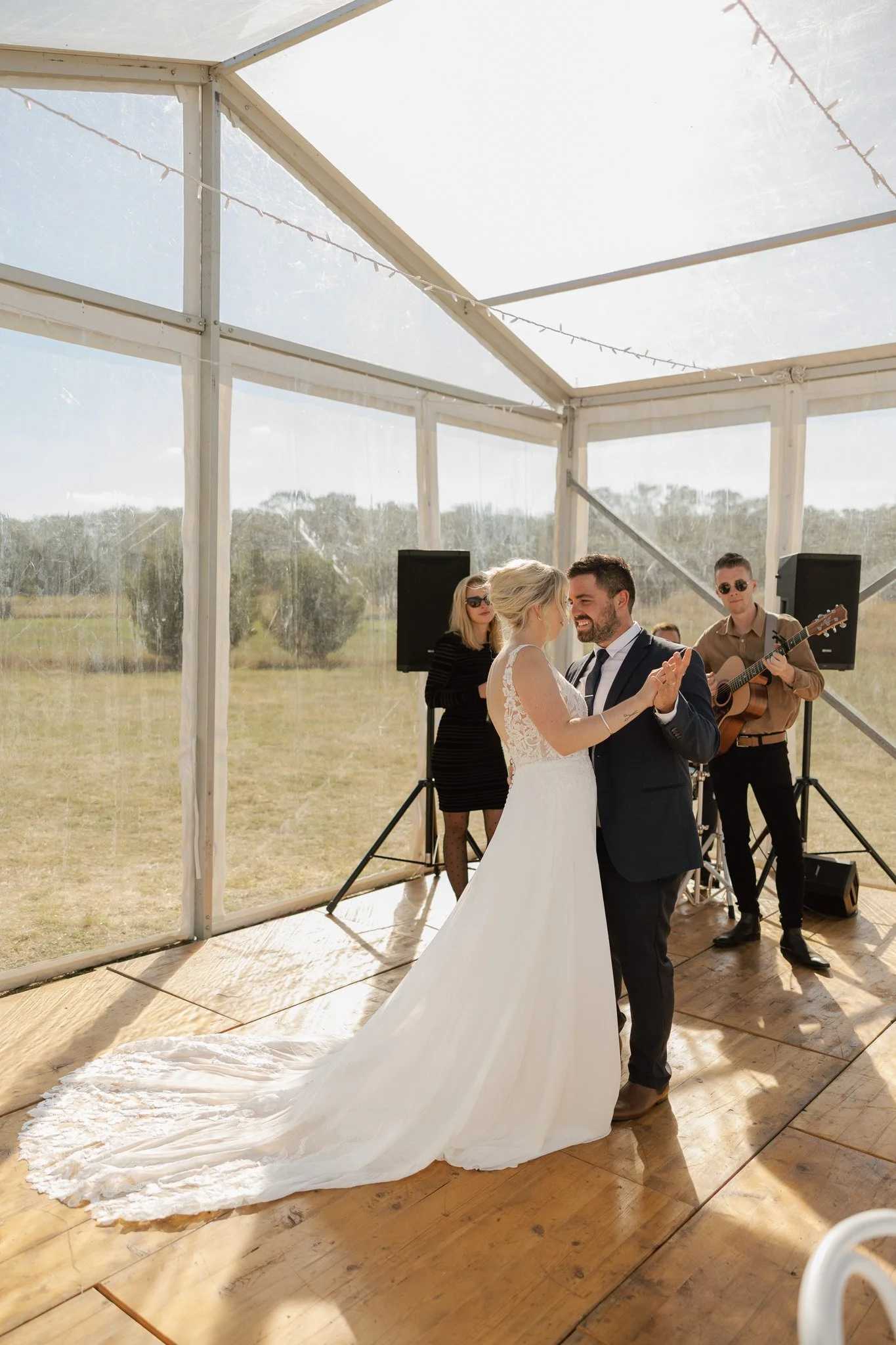 A bride and groom are dancing at their wedding reception inside a transparent tent, with a musician playing guitar in the background.